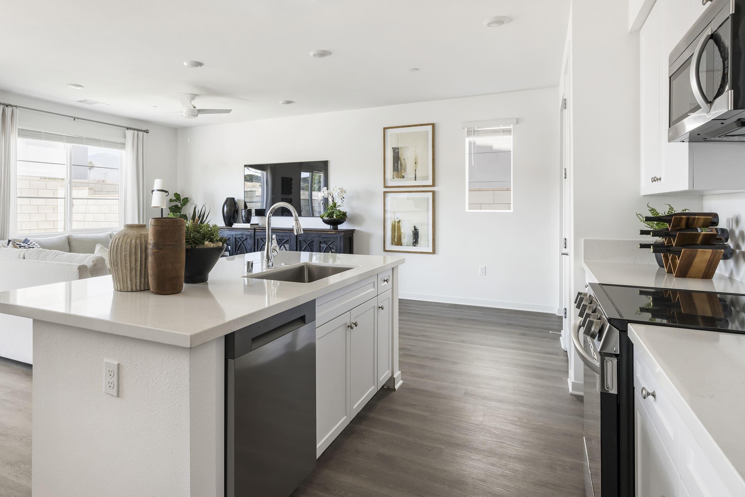 Modern open-concept kitchen and living area featuring a sleek island with a sink, stainless steel appliances, and a dishwasher. The living room includes a comfortable sofa, a television, and decorative artwork on the walls. Large windows provide ample natural light and a view of the exterior.