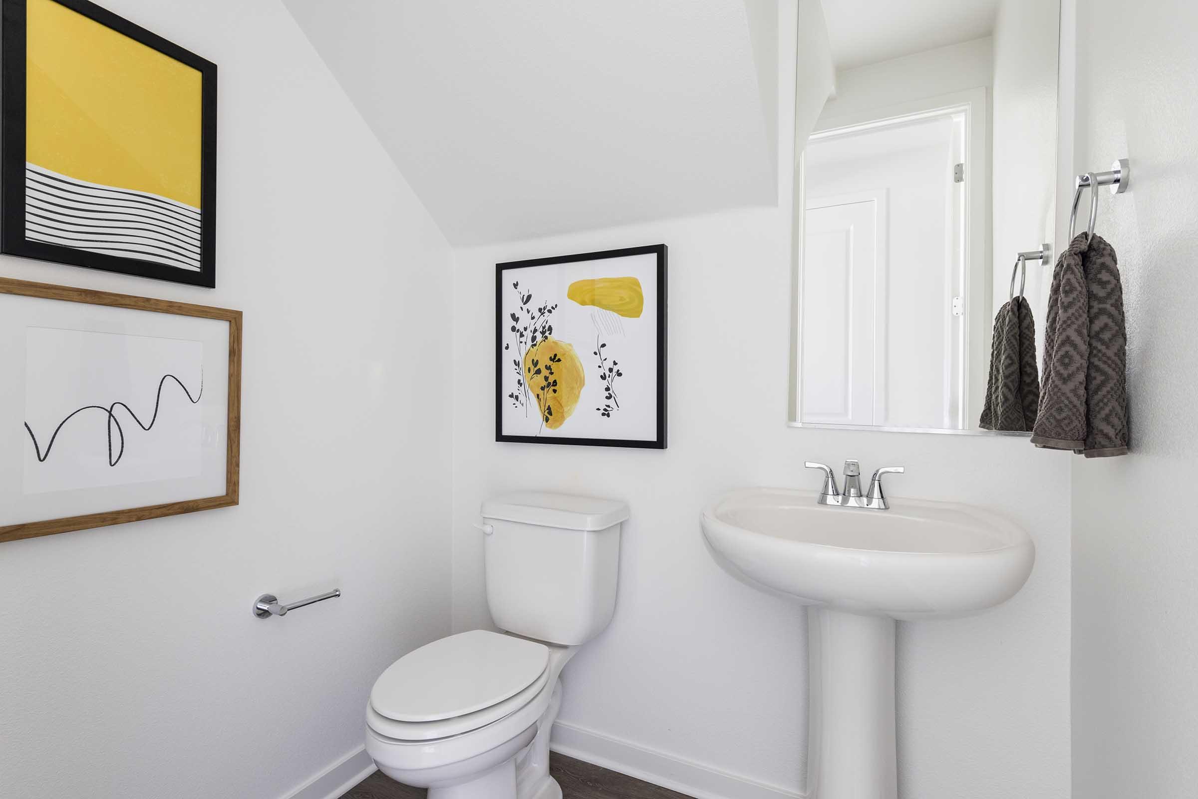A modern bathroom featuring a white wall, a toilet, and a pedestal sink. Two framed art pieces are displayed on the wall, one with a yellow and black design and the other with abstract shapes. A small mirror reflects the room, and two hand towels hang on a rack.