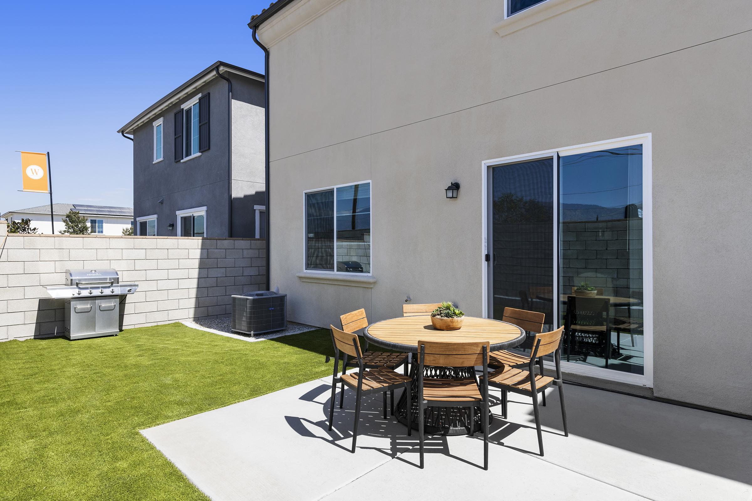 A modern outdoor patio featuring a round wooden table surrounded by six chairs, with a potted plant on the table. To one side, there is a barbecue grill and a grassy area, while a beige house with large windows and a flag is in the background under a clear blue sky.
