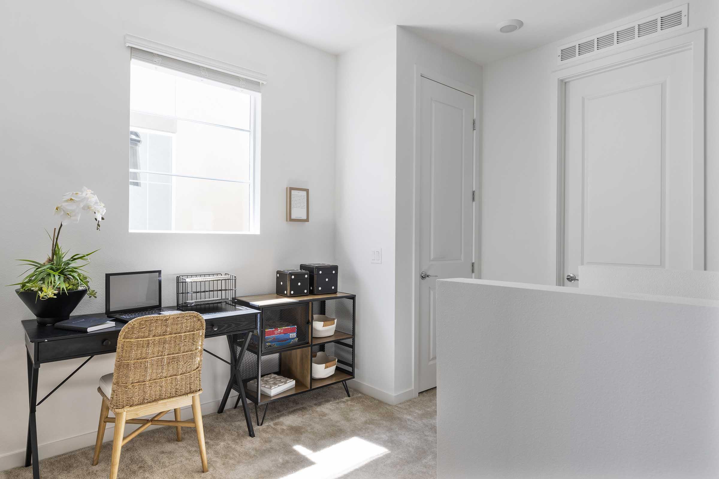 A bright, modern home office space featuring a small black desk with a laptop, a decorative plant, and a storage basket. The room has white walls, a window allowing natural light, a closed door, and a shelf with various storage boxes and items. Soft beige carpet covers the floor.