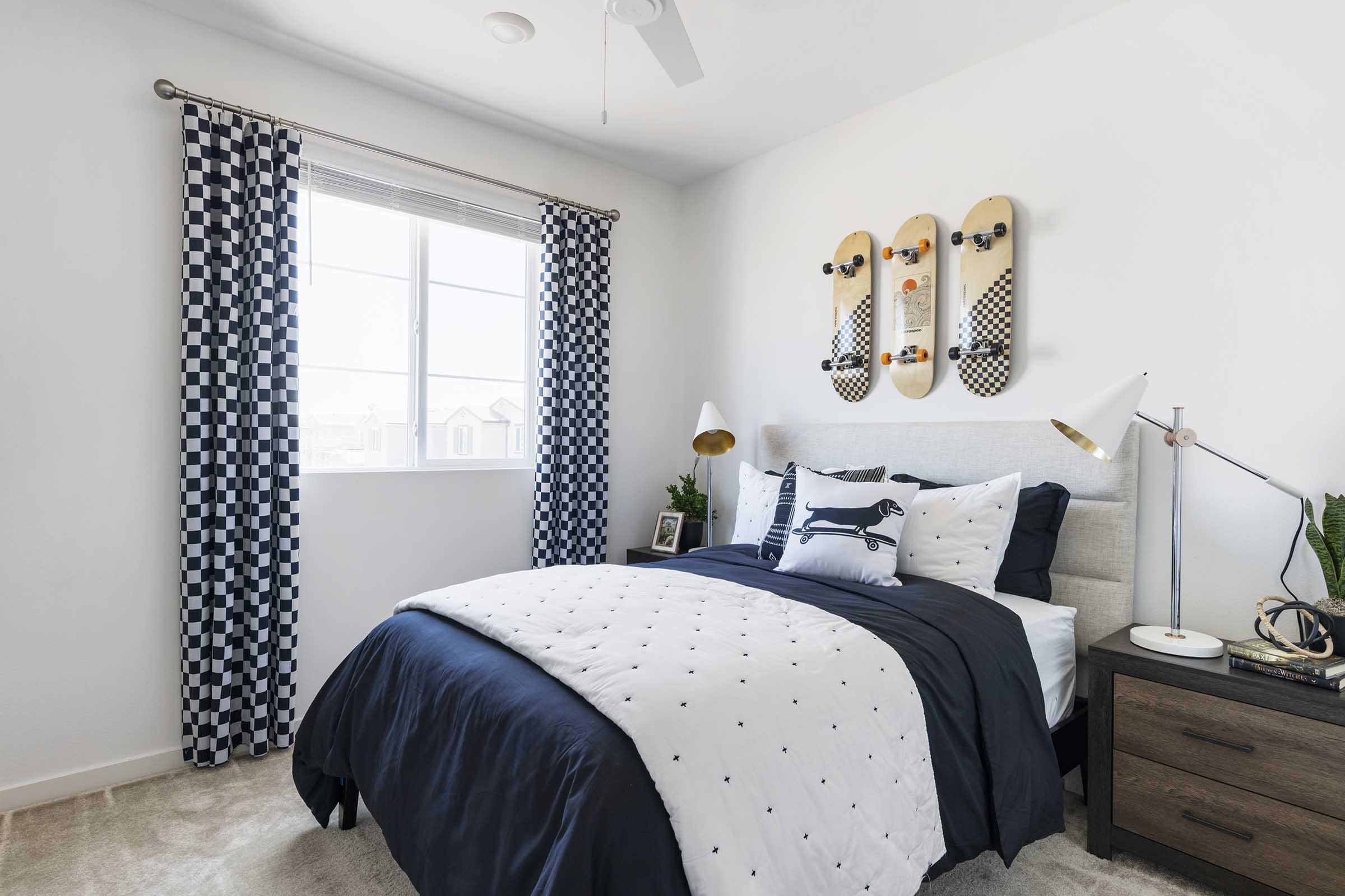 A stylish bedroom featuring a queen-sized bed with white and navy bedding, accented by decorative pillows. There are checkered curtains, a wooden nightstand with a plant and a picture frame, and three skateboard decor pieces on the wall above the bed. Natural light comes through a window, enhancing the modern aesthetic.