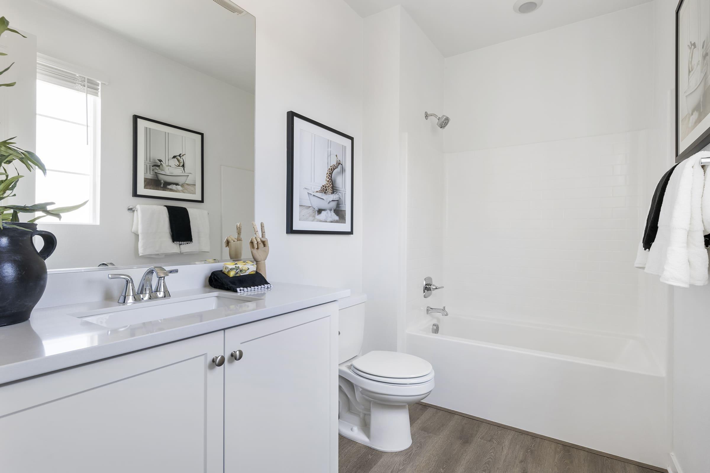 A modern bathroom featuring a white bathtub and shower combination, a sleek vanity with a mirror, and minimalist decor. There are framed art pieces on the wall, a black vase with plants, and neatly arranged towels. The overall design is bright and airy with light-colored walls and wooden flooring.