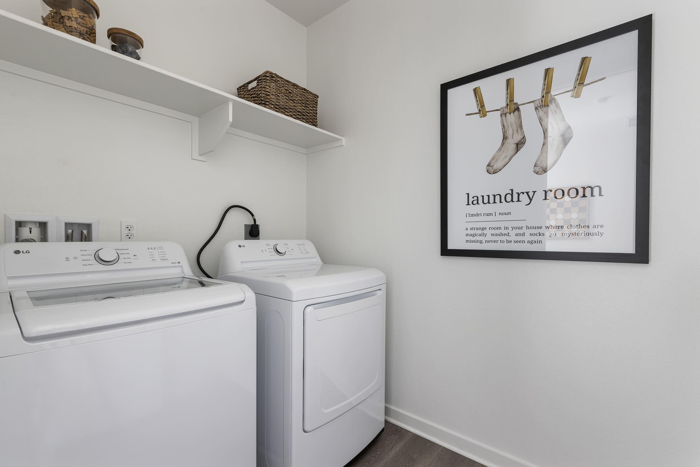 A laundry room featuring a washing machine and a dryer side by side. There's a shelf above with a woven basket, and a framed poster on the wall displaying socks hanging from clothespins with the text "laundry room." The walls are painted white, and the flooring is light-colored.