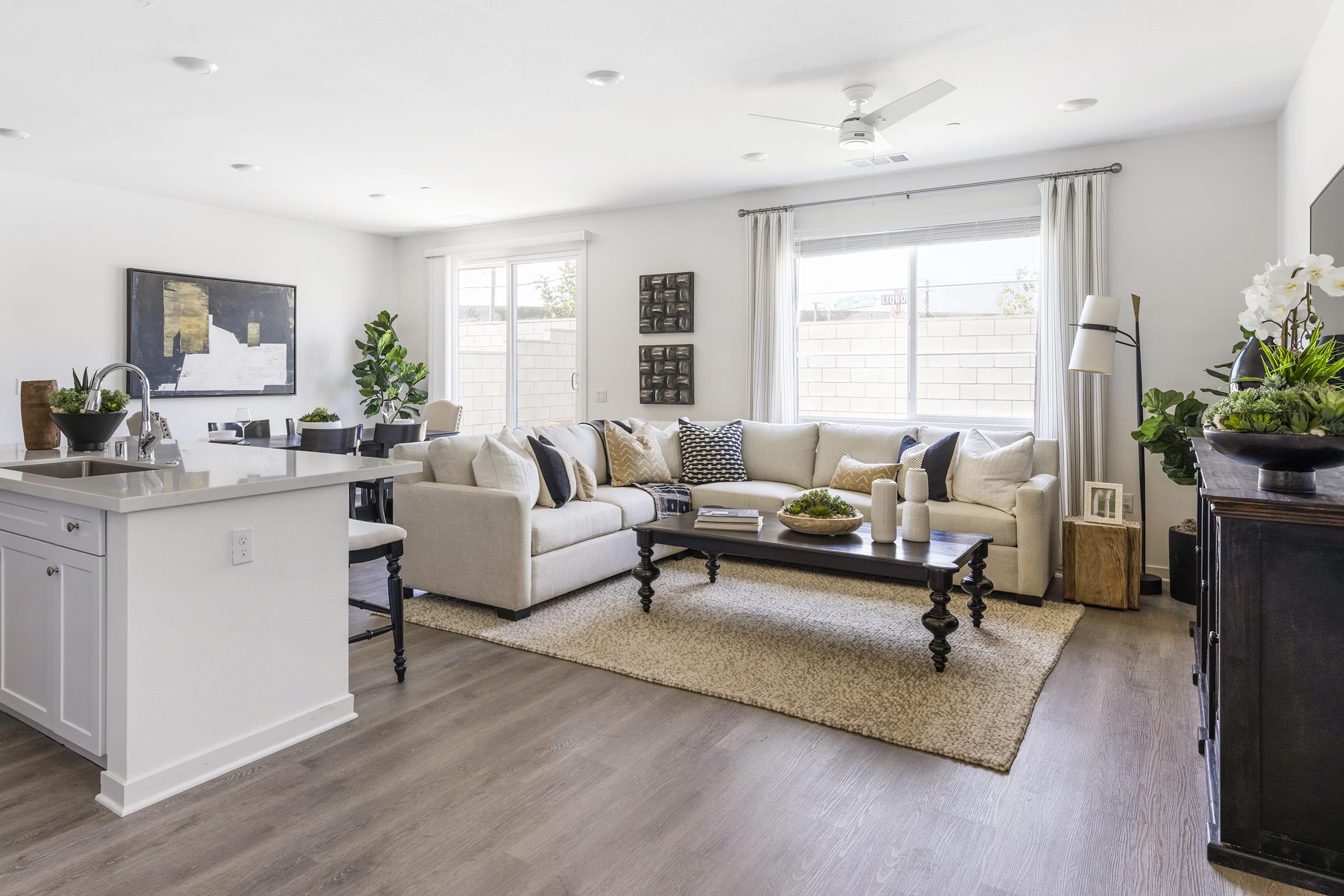 Bright and modern living room featuring a comfortable beige sectional sofa with decorative pillows, a wooden coffee table, and a large window allowing natural light. The space includes greenery, a stylish rug, and a dining area with sleek cabinetry, creating a cozy and inviting atmosphere.