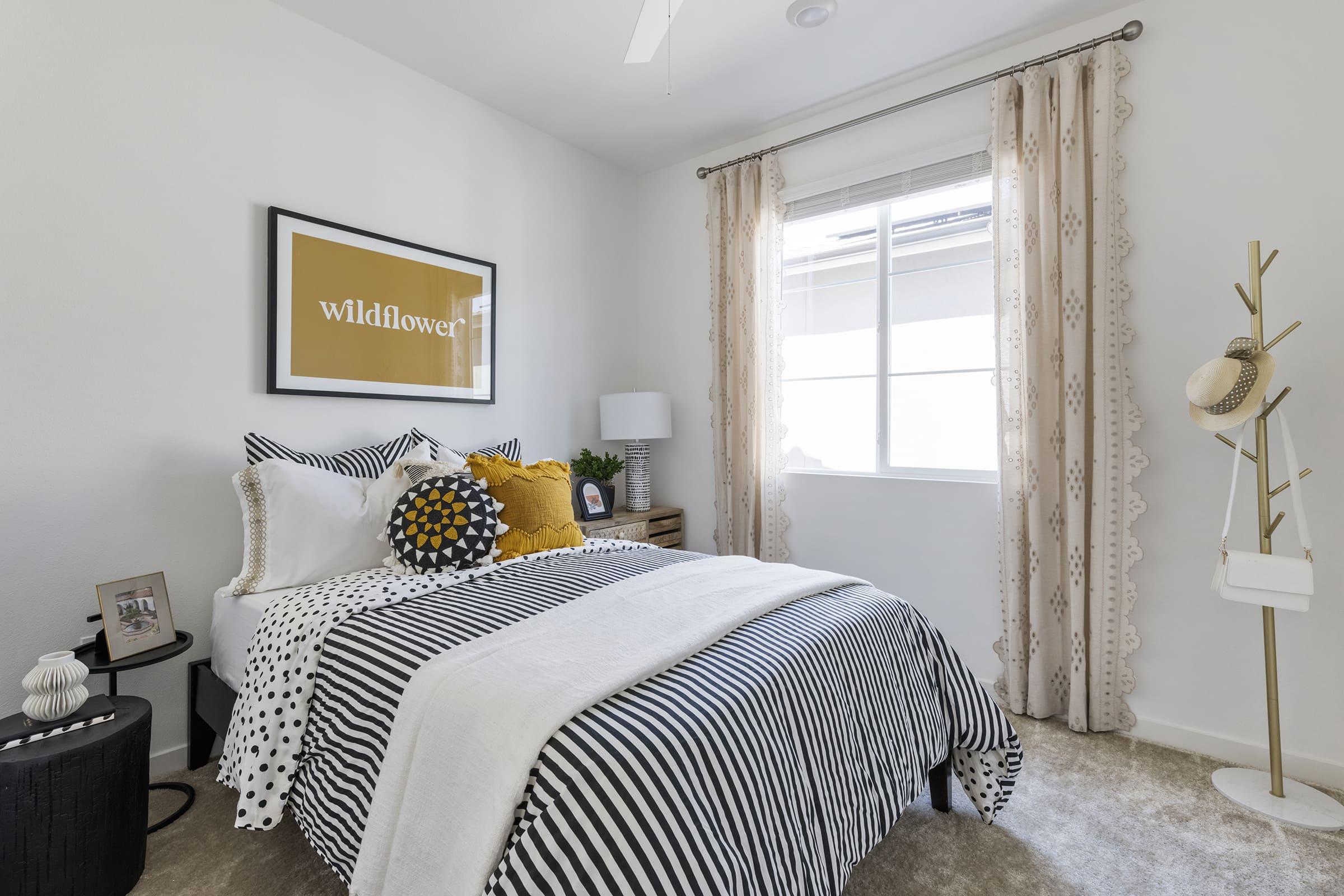 A cozy bedroom featuring a black and white striped bedspread with a mix of patterned pillows. There's a beige throw blanket on the bed, a nightstand with a lamp, and an art piece above the bed that reads "wildflower." A window allows natural light in, and a decorative coat rack stands in the corner.