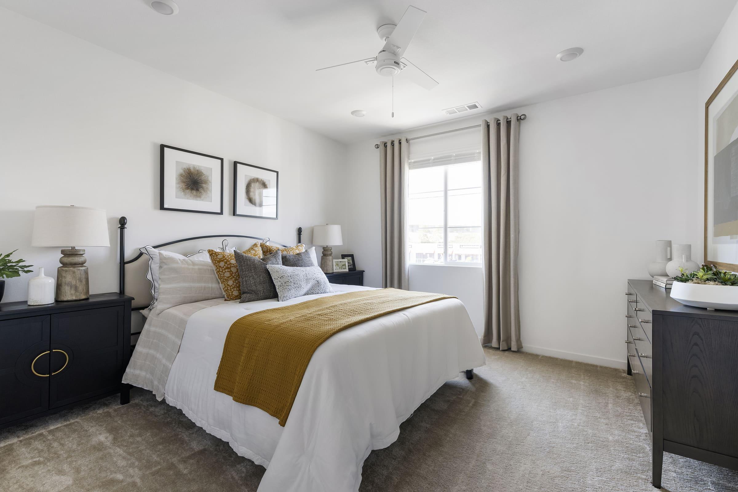 A modern bedroom featuring a queen-sized bed with a white comforter and mustard yellow throw blanket. The walls are light, with framed art pieces above the bed. A bedside table with a lamp is on one side, and a wooden dresser is on the opposite side. Natural light floods the room through a window with sheer curtains.