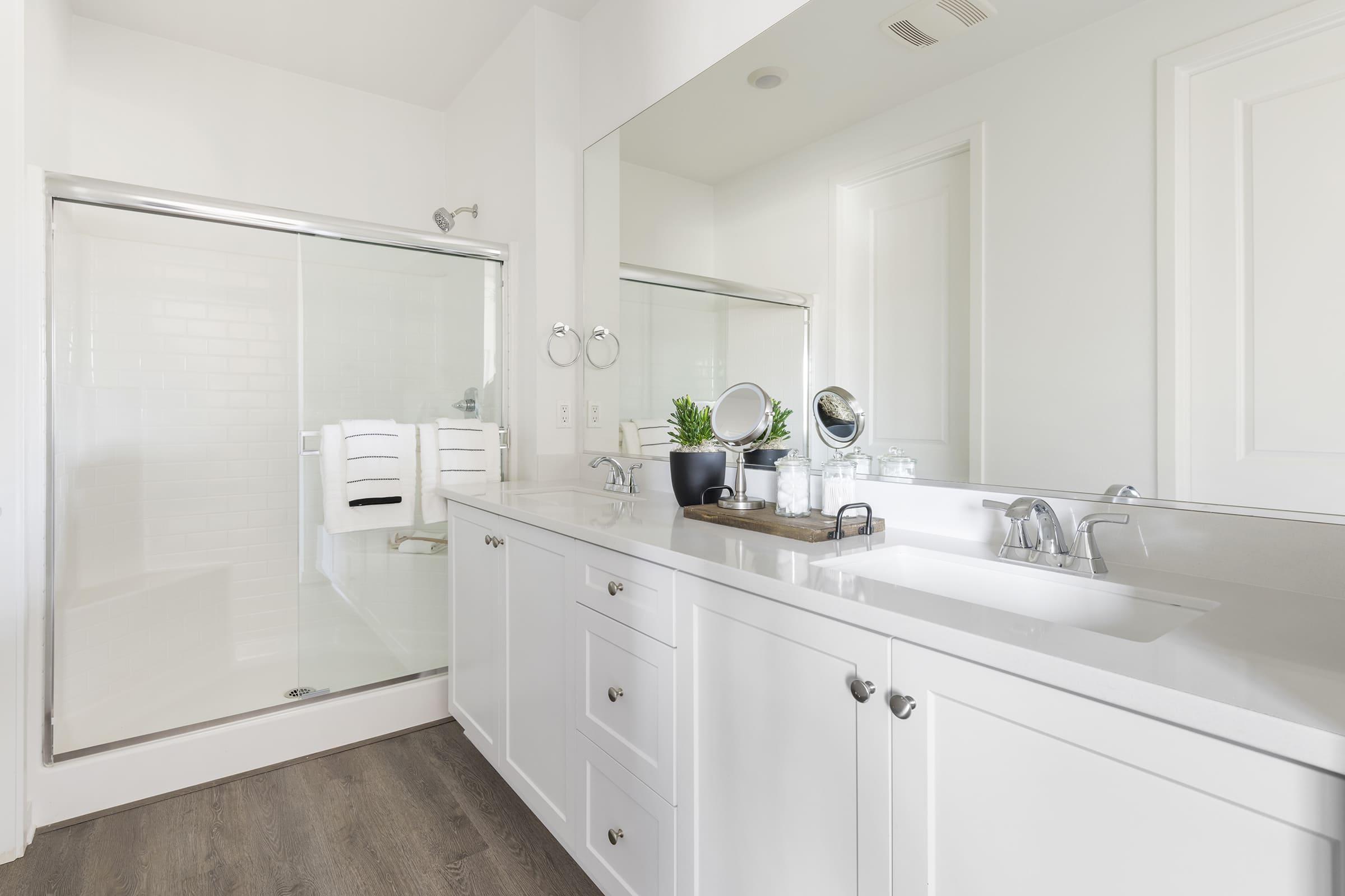 A modern bathroom featuring a white vanity with storage drawers, a double sink, and a large mirror. To the left, there is a glass shower enclosure with white towels hanging. The decor includes a small plant and a round mirror on the vanity, set against light-colored walls and a wood-look floor.