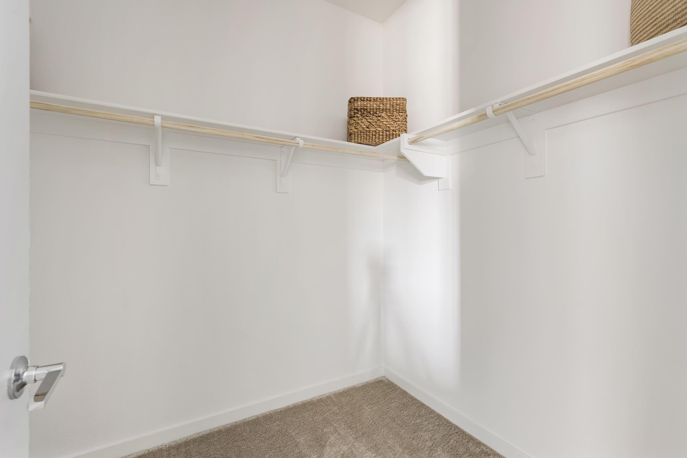 Empty walk-in closet with white walls and a light tan carpet. Two wooden shelves are mounted on the walls, and a woven basket sits on the top shelf. A door is slightly ajar, revealing the closet’s interior, which is bright and spacious.