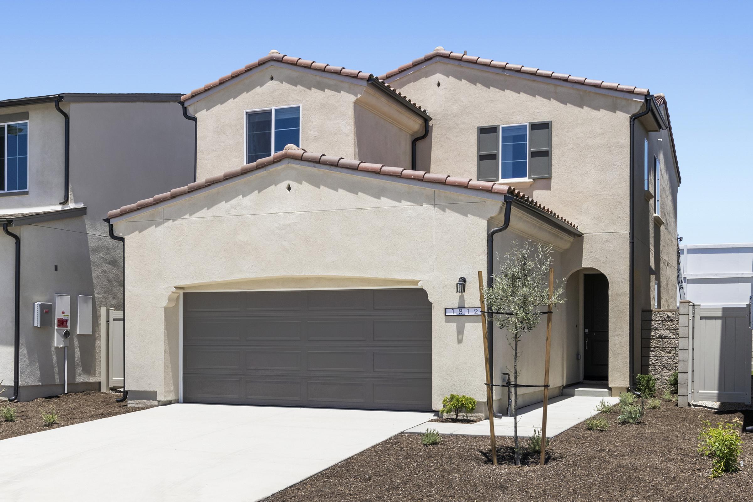 Two-story modern house with a stucco exterior and a tiled roof. The front features a two-car garage, landscaped yard with small plants, and large windows. The driveway is paved, and the surrounding area is neatly organized, showcasing a contemporary architectural style.