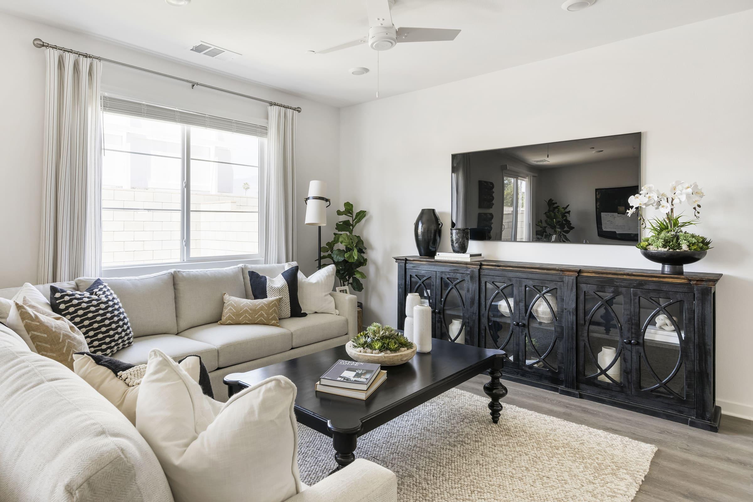 A modern living room featuring a light gray sectional sofa with decorative pillows, a black coffee table, and a stylish black console beneath a large flat-screen TV. There are green plants, a white vase with flowers, and a textured rug on the floor, creating a cozy and elegant atmosphere.