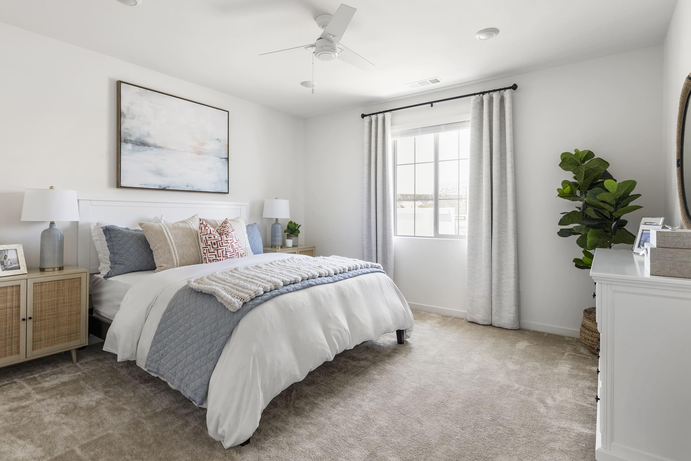 A softly decorated bedroom featuring a neatly made bed with a light blue and white comforter. There are two bedside tables with lamps, a large artwork on the wall, and a potted plant in the corner. Natural light streams in through a window with white curtains, creating a serene and inviting atmosphere.
