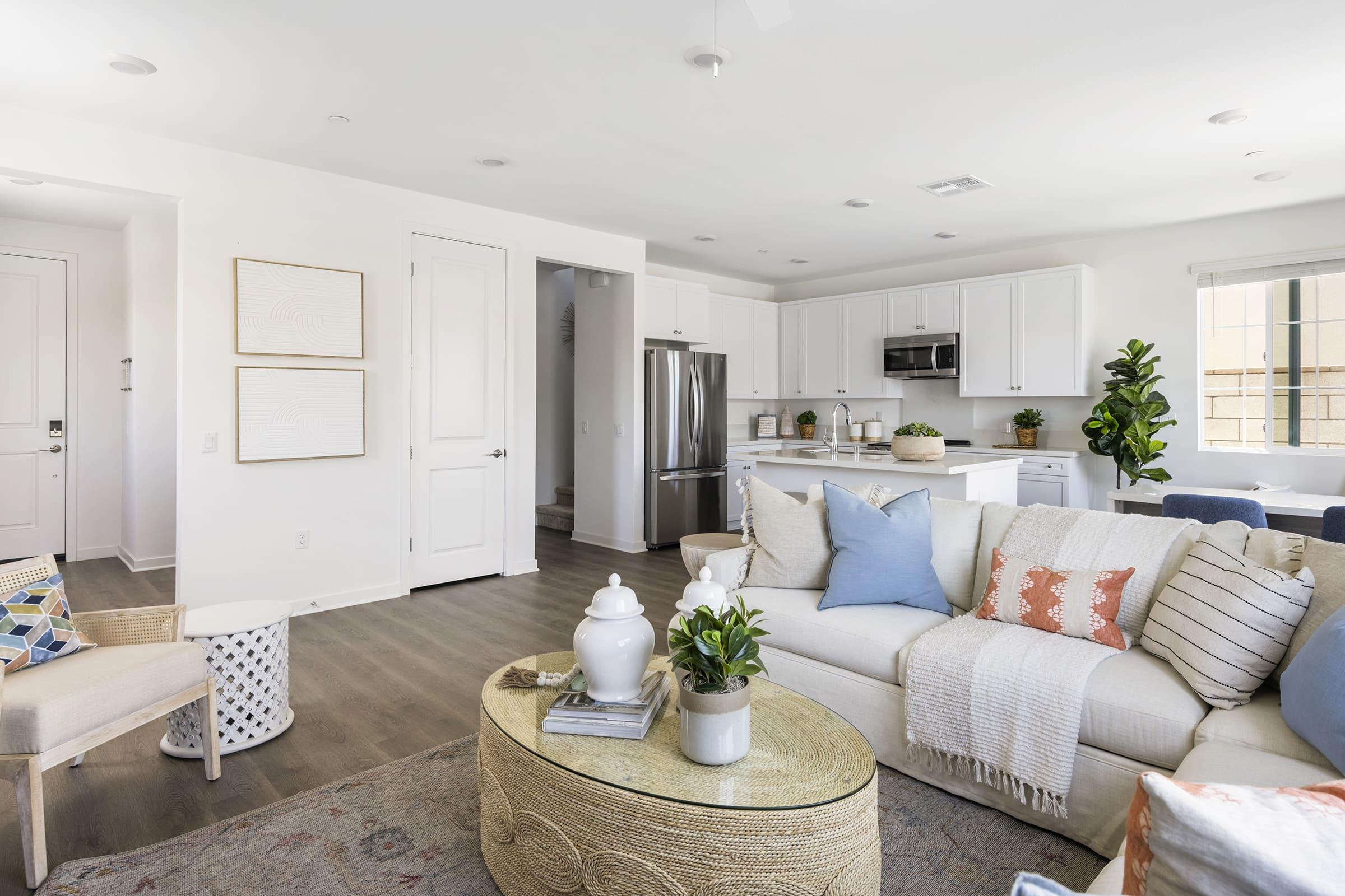 A bright and modern living room featuring a spacious sofa adorned with various decorative pillows, a round coffee table, and a cozy armchair. The kitchen area is visible in the background with white cabinets and stainless steel appliances, complemented by large windows that let in natural light.