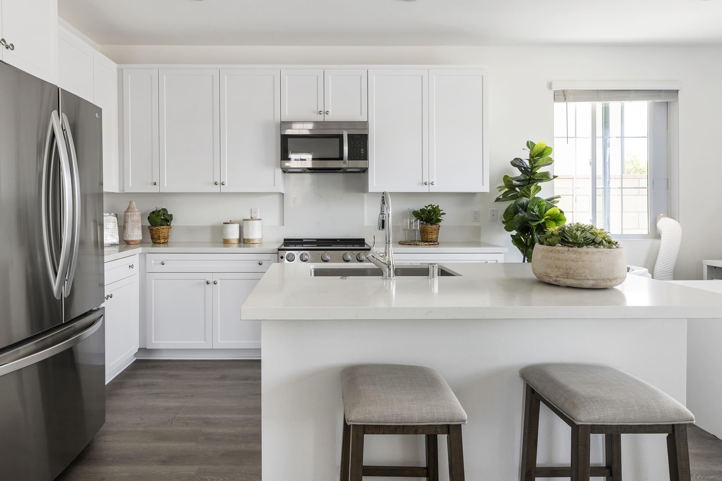 Modern kitchen featuring white cabinetry, stainless steel appliances, and a large island with bar stools. A potted plant and decorative items add a touch of greenery. Natural light streams in through a window, highlighting the sleek design and open layout.