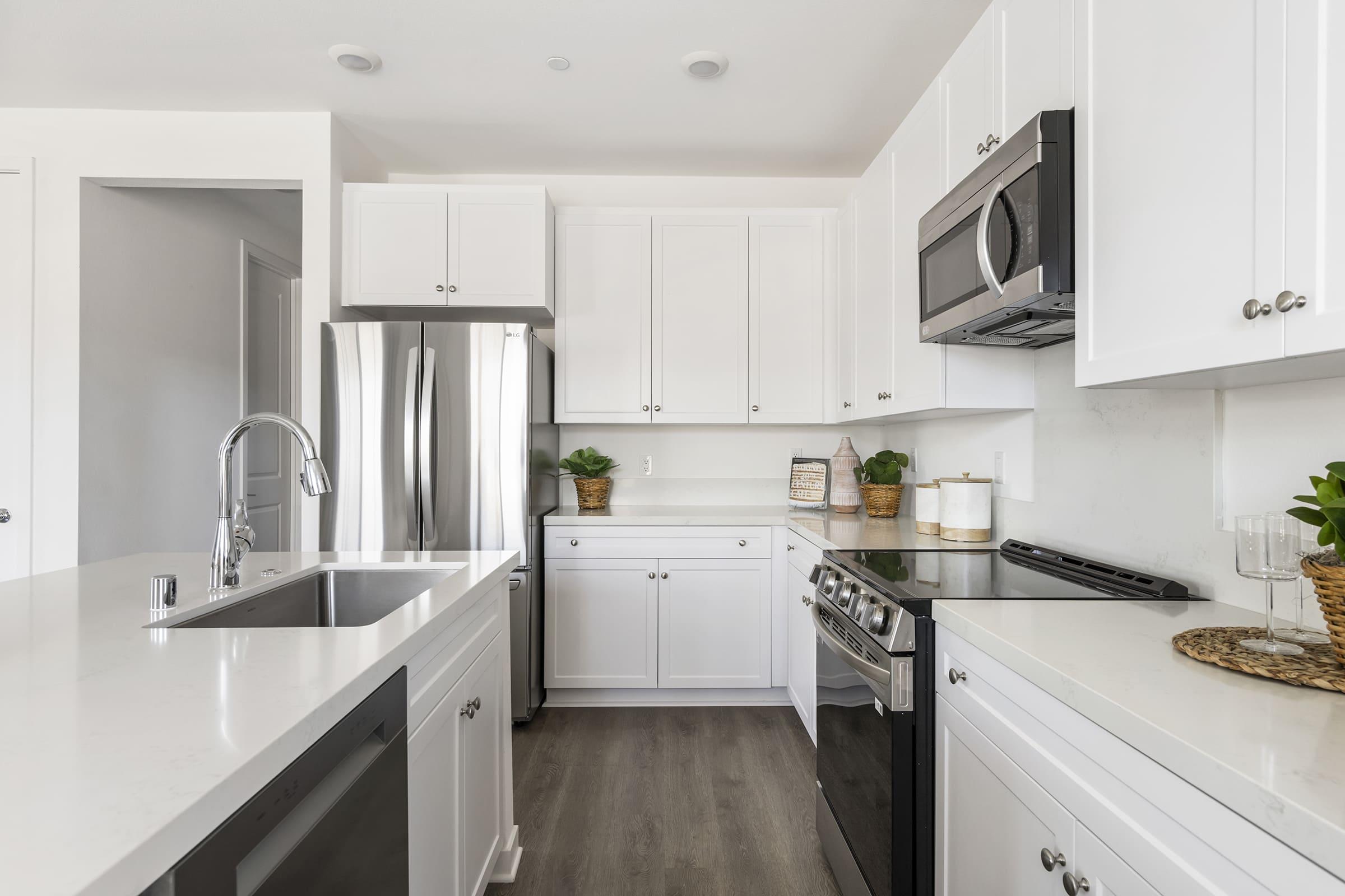 A modern kitchen featuring white cabinetry, stainless steel appliances including a refrigerator and oven, and a sleek countertop. The space is well-lit, with plants and decorative items on display, creating a clean and inviting atmosphere.