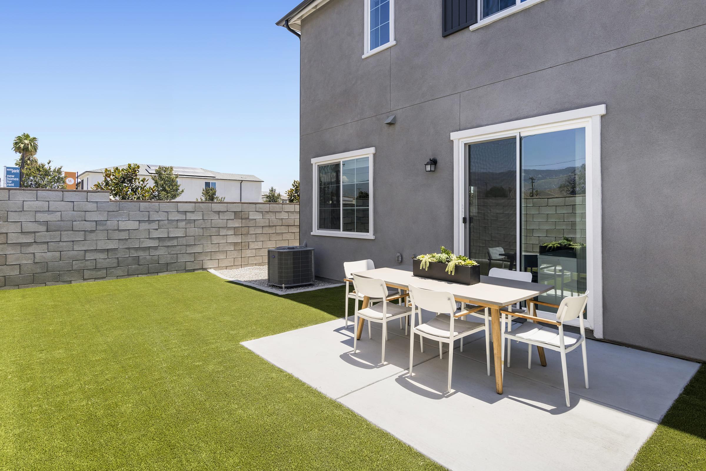 A modern backyard with artificial grass, featuring a dining table set with white chairs. The house has a grey exterior, large windows, and a sliding glass door. There’s an air conditioning unit on the side and a simple stone wall in the background, under a clear blue sky.