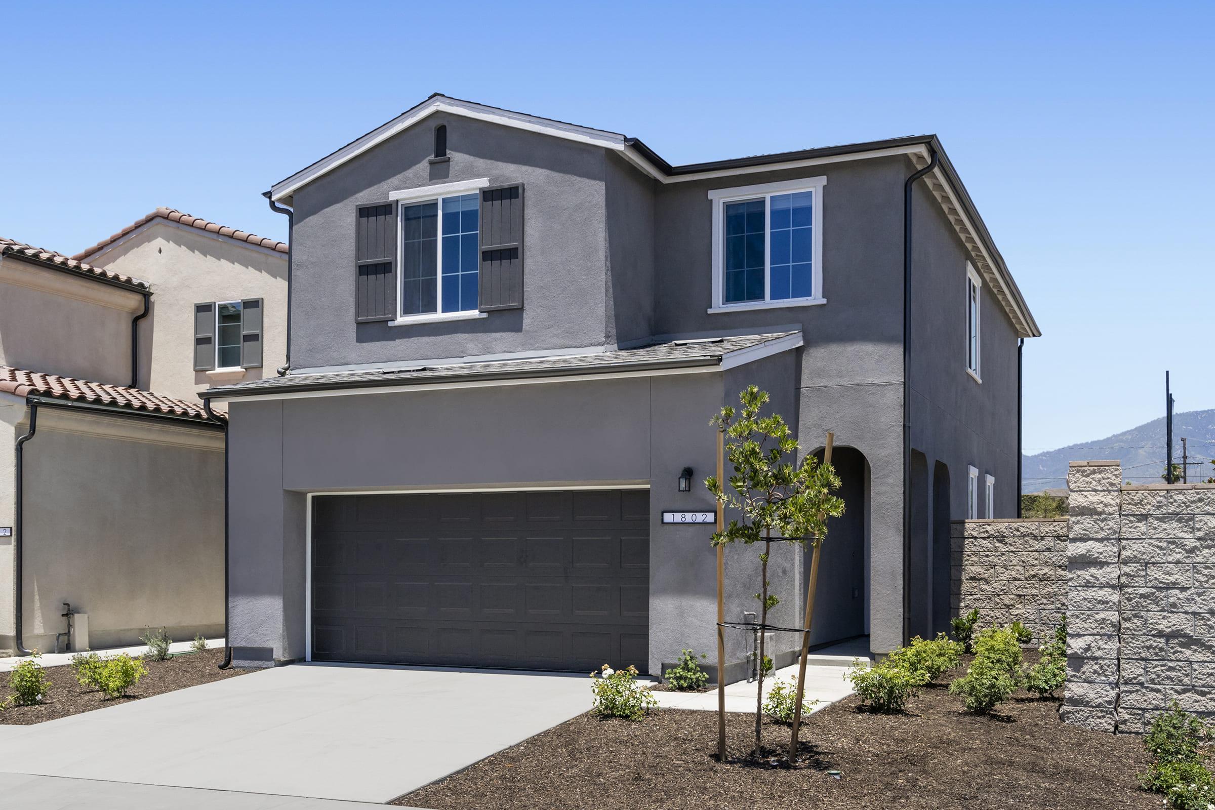 A modern two-story house with gray exterior, black shutters, and a two-car garage. The front yard features low-maintenance landscaping with small shrubs and a concrete driveway. The setting is sunny, with a clear blue sky and distant hills visible in the background.
