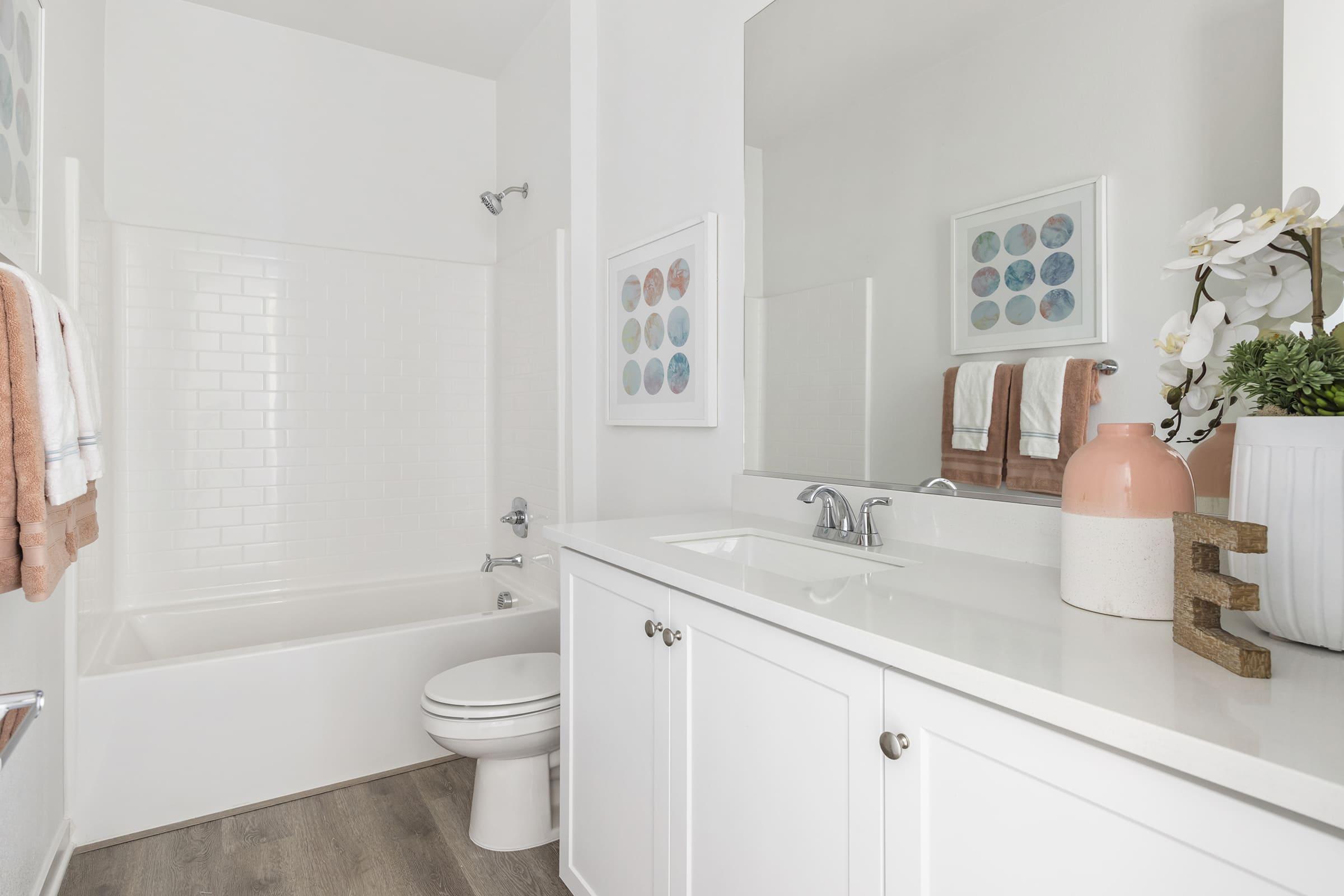 A bright and modern bathroom featuring a white bathtub, a clean vanity with a sink, and a large mirror. Soft-colored towels hang on a rack, and decorative elements include a potted plant, artwork with circular patterns, and a neutral-toned vase. The overall design is fresh and inviting.