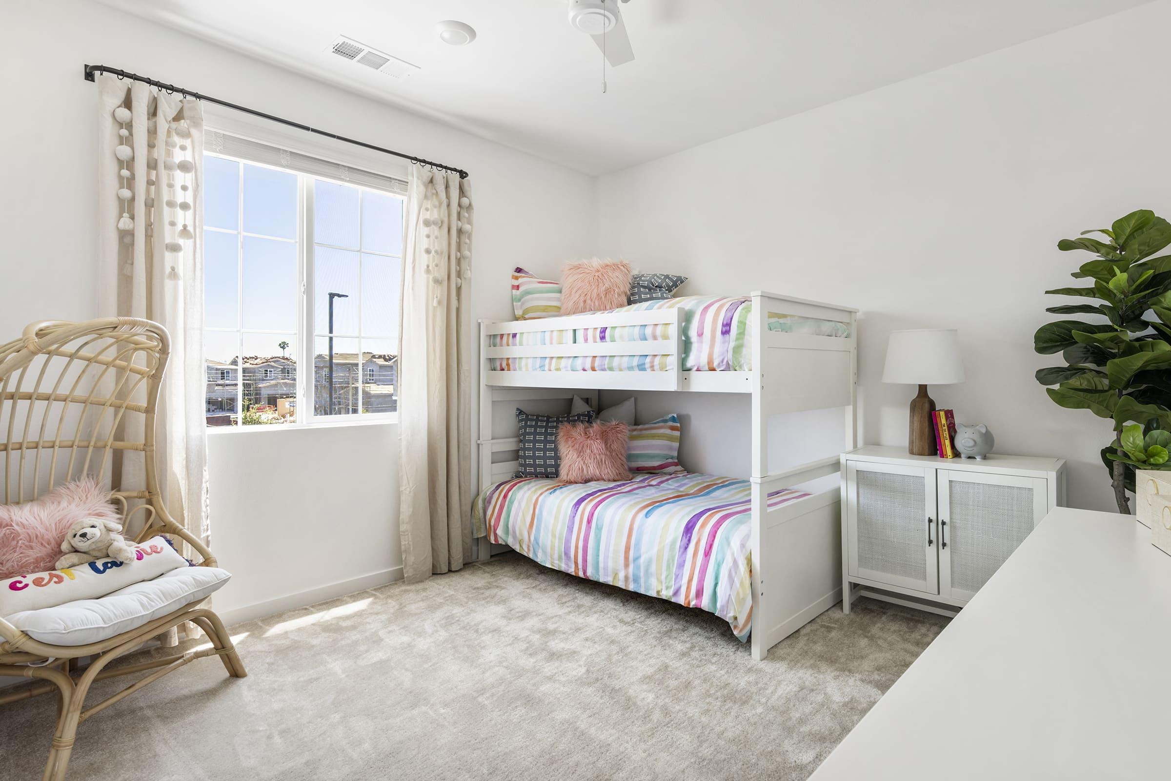 A bright, airy children's bedroom featuring a bunk bed with colorful striped bedding. A cozy rattan chair with decorative pillows sits in one corner, and a stylish white dresser is against the wall. Large windows let in natural light, and a green plant adds a touch of nature to the room.