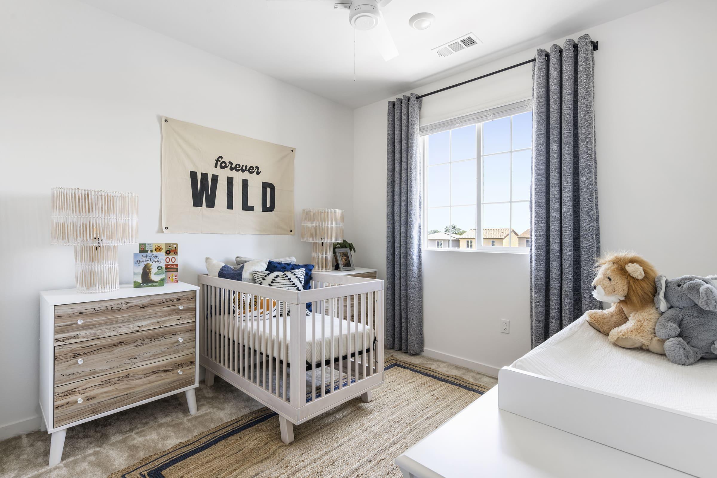 A bright and airy nursery featuring a white crib with a "forever WILD" wall hanging above it. The room has gray curtains, a patterned area rug, and two decorative lamps on a wooden dresser. There's a stuffed lion on the dresser and a gray stuffed animal in the crib, suggesting a playful, nature-inspired theme.