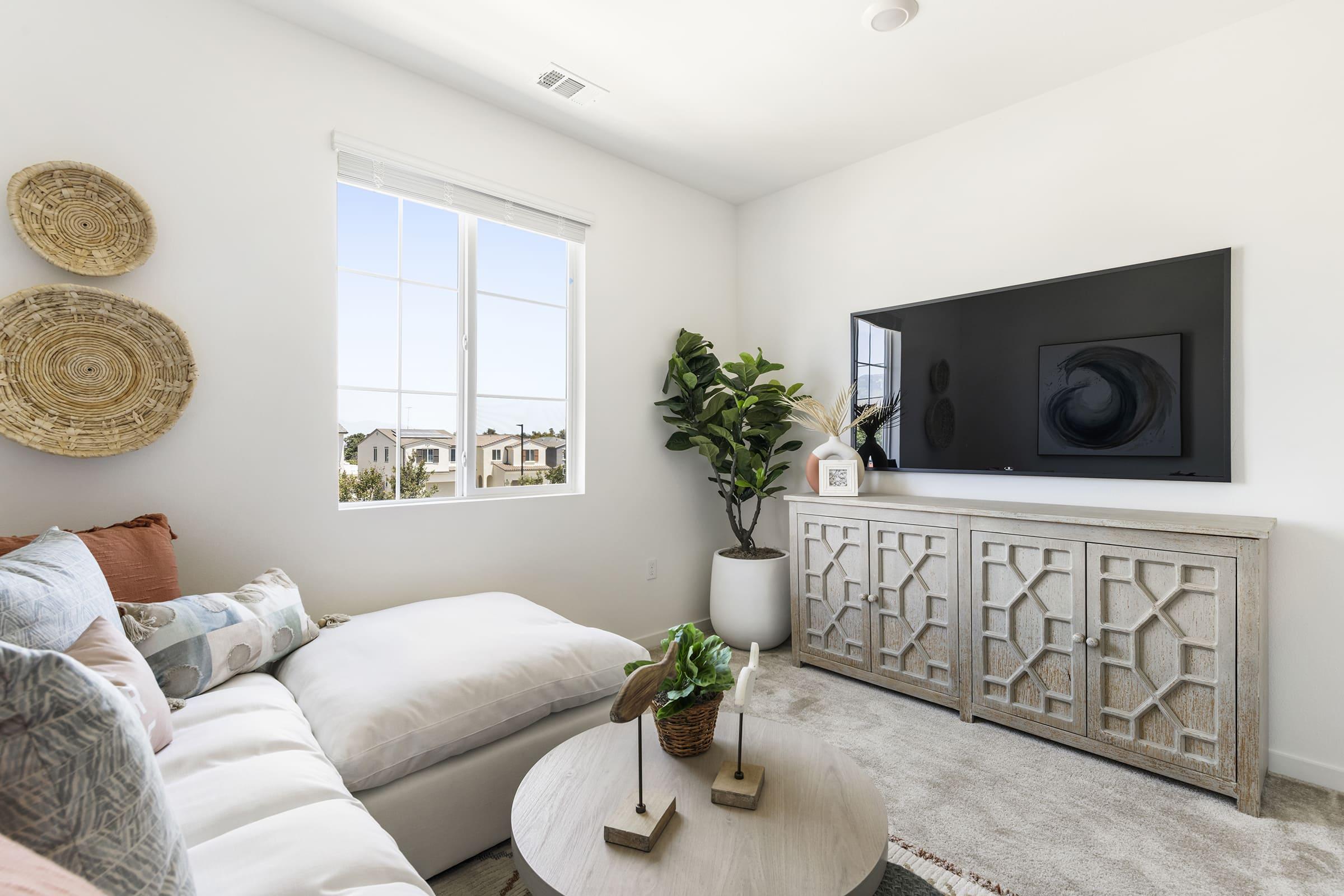 A cozy living room featuring a large window with natural light, a sectional sofa adorned with colorful pillows, a modern TV mounted on the wall, and a stylish wooden cabinet. A potted plant and decorative items add a fresh touch to the space, creating a warm and inviting atmosphere.