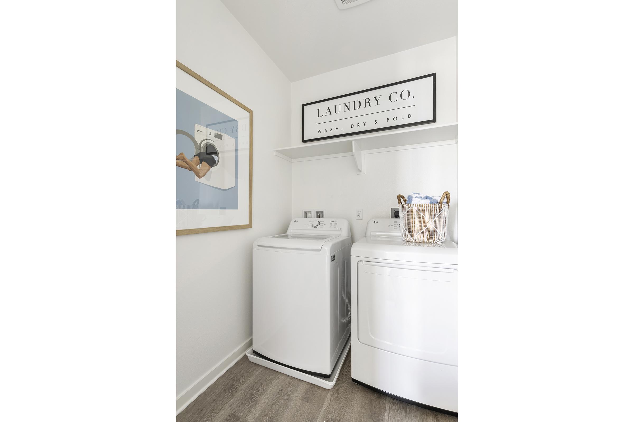 A tidy laundry room featuring a white washing machine and dryer side by side. Above them, a decorative sign reading "LAUNDRY CO." is mounted on the wall. A framed artwork is hung to the left, and a basket is placed on the dryer. The space has light-colored walls and wooden flooring, creating a bright atmosphere.
