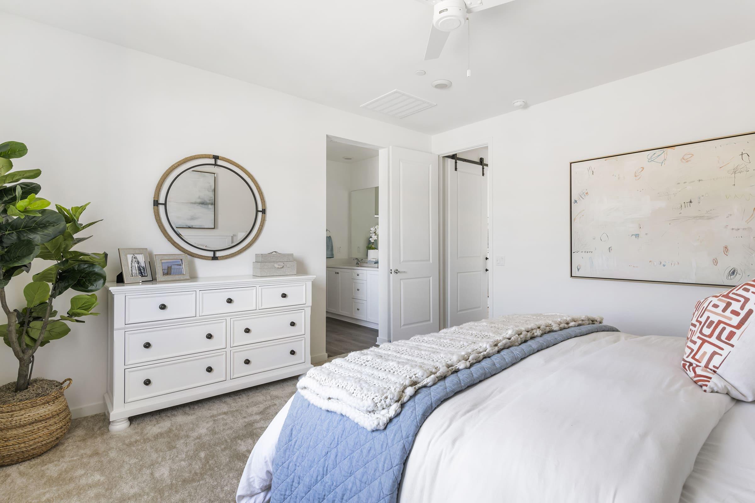 A modern bedroom featuring a neatly made bed with a blue throw blanket. A white dresser with photographs and a circular mirror adorn the wall. In the background, a bathroom is partially visible through an open door. A potted plant adds a touch of greenery to the room. The decor is light and inviting.