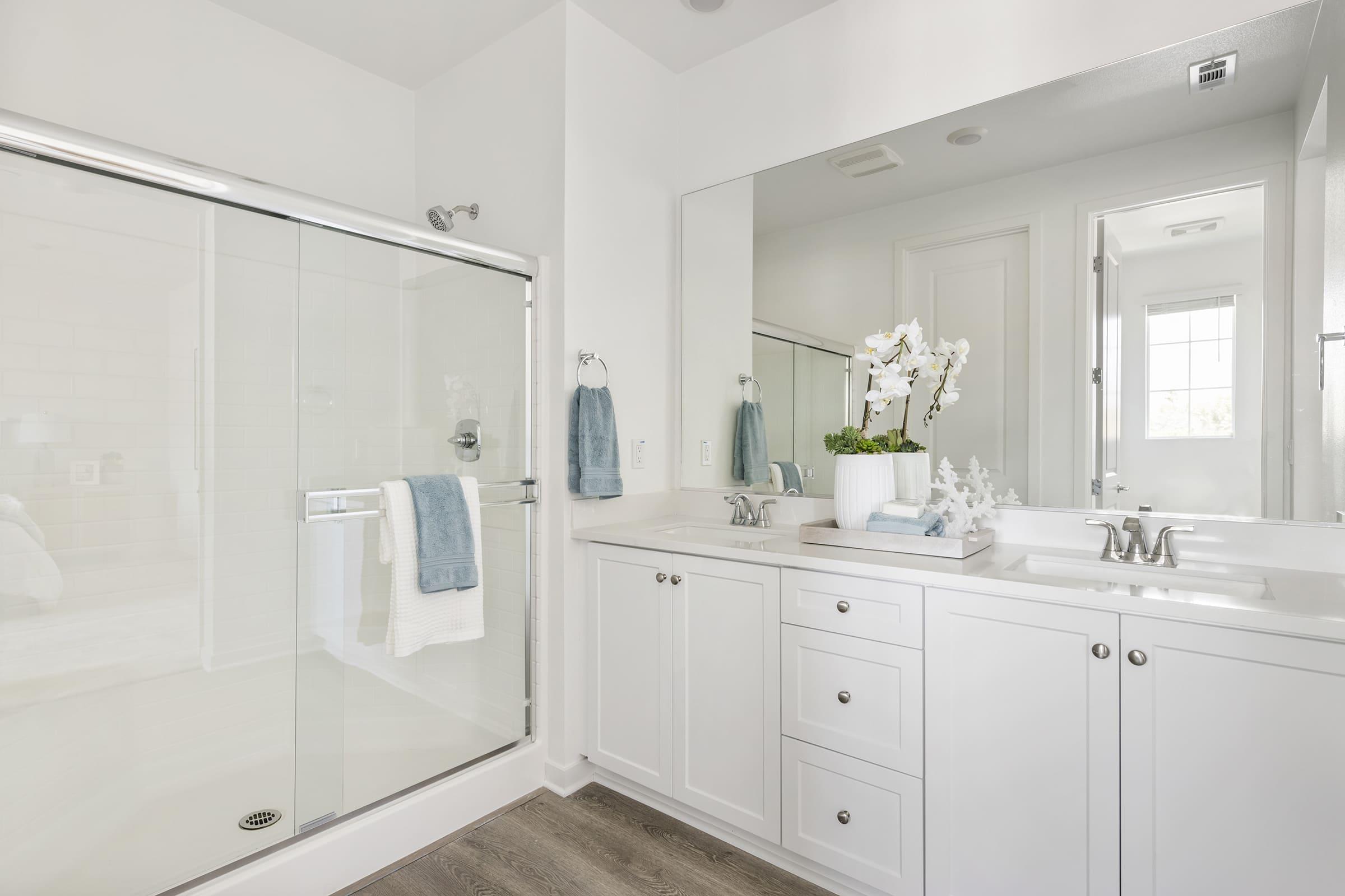 A modern bathroom featuring a glass shower enclosure, a white vanity with drawers, and a large mirror. The decor includes white towels and a stylish vase with an orchid plant. Natural light streams in through a nearby window, enhancing the fresh, clean aesthetic.