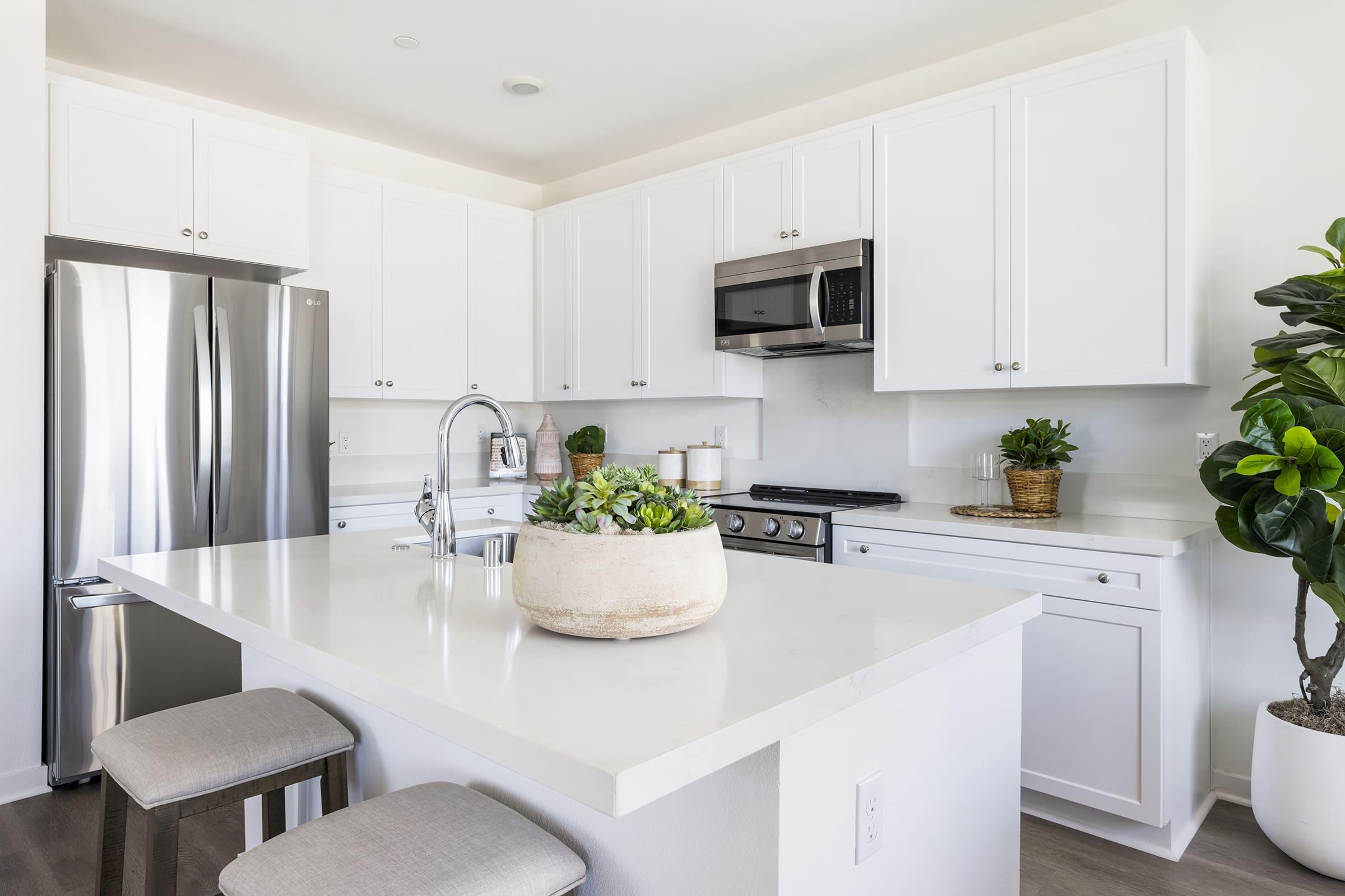 A modern kitchen featuring white cabinetry, stainless steel appliances, and a sleek countertop. The kitchen island has a large planter with greenery, and there are two beige bar stools at the island. Natural light illuminates the space, creating a bright and inviting atmosphere.