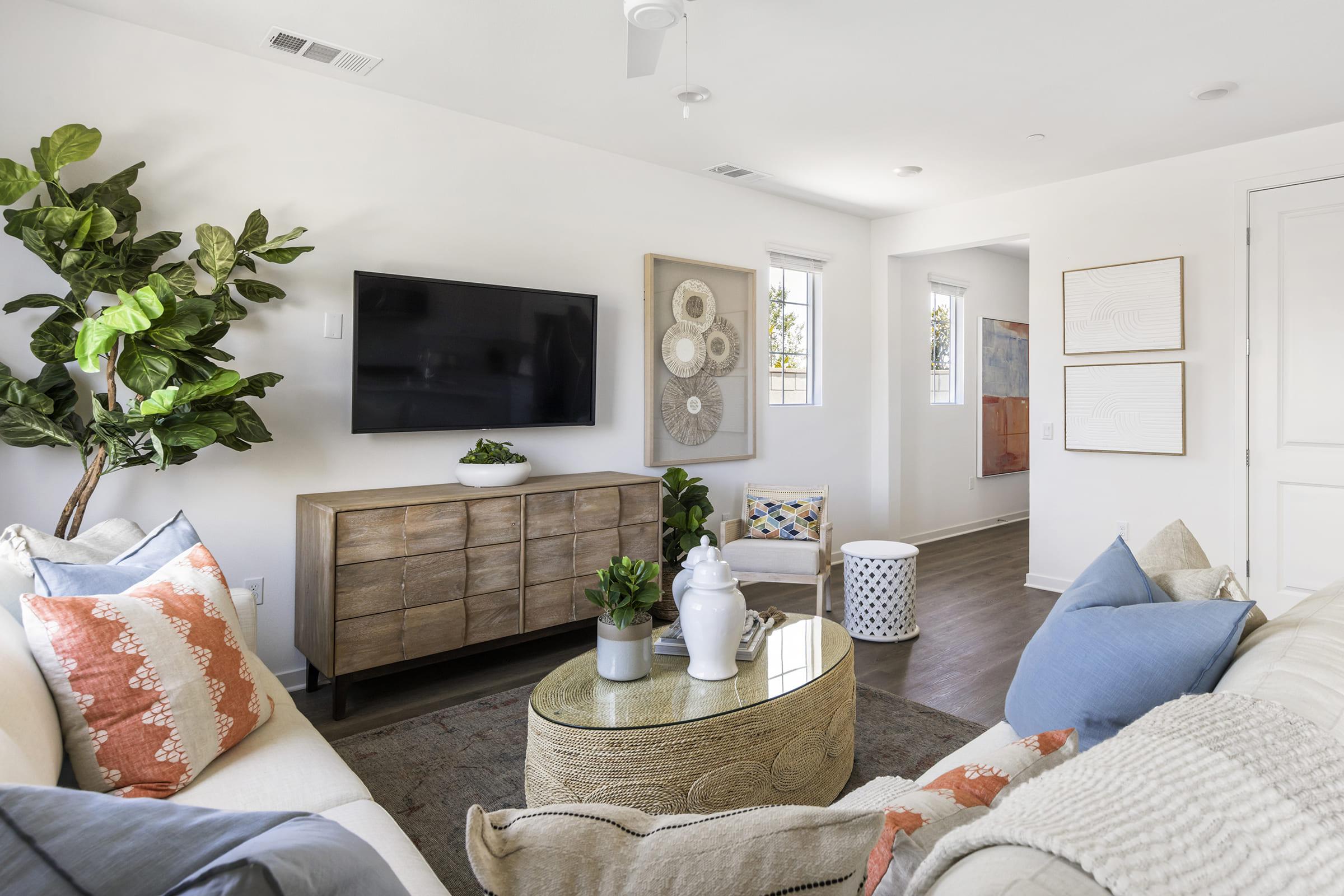 A modern living room featuring a comfortable cream-colored sofa adorned with blue and coral pillows. A round coffee table sits in front of the sofa, topped with a decorative white vase and greenery. A large TV is mounted on the wall, and a wooden sideboard offers additional decor. Large windows invite natural light.
