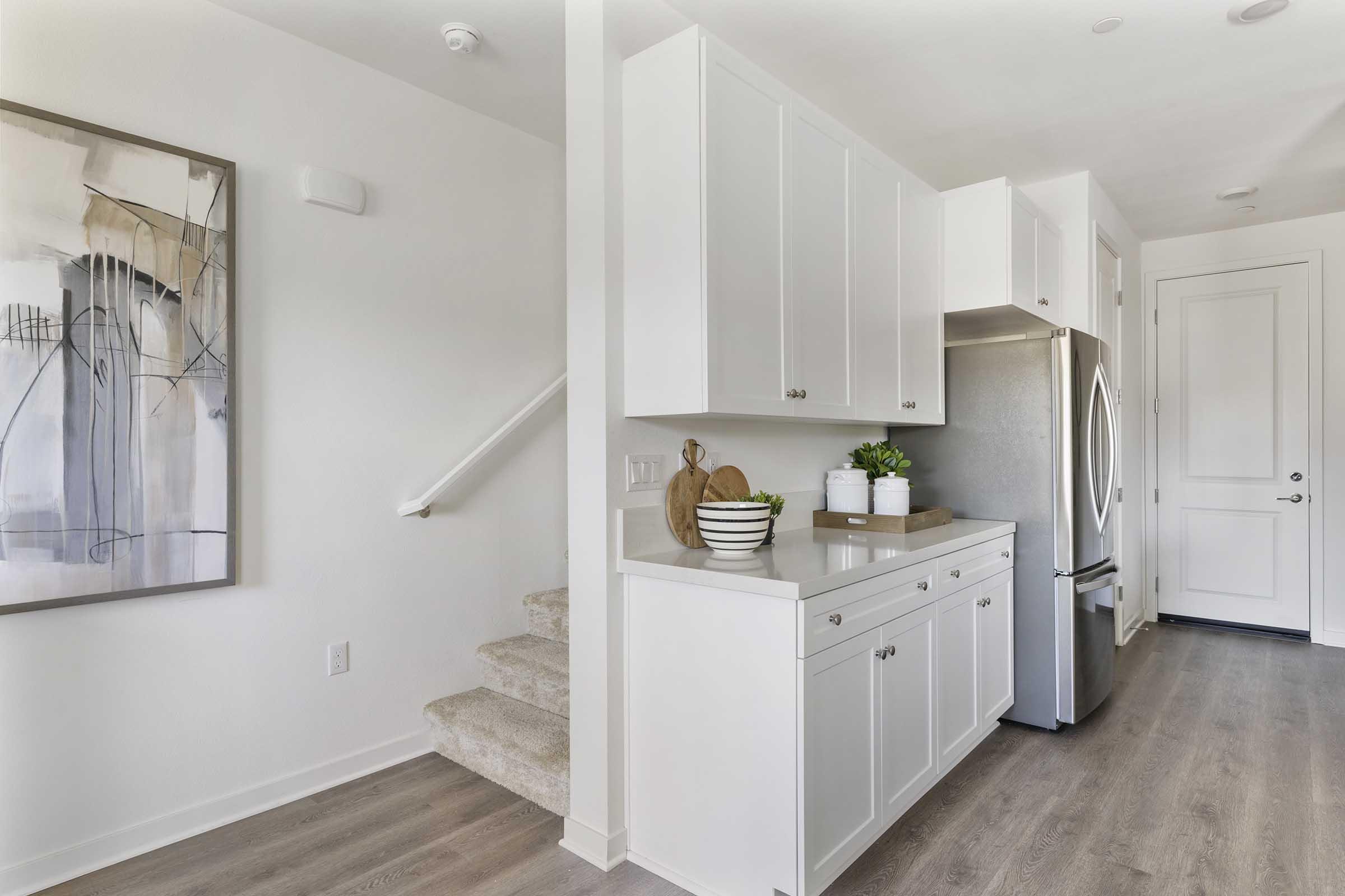 A modern kitchen featuring white cabinetry, a stainless steel refrigerator, and decorative items on the countertop. A staircase is visible on the left, and a large abstract painting adorns the wall, adding a touch of elegance to the space. The floor has light wood tones, creating a bright, inviting atmosphere.