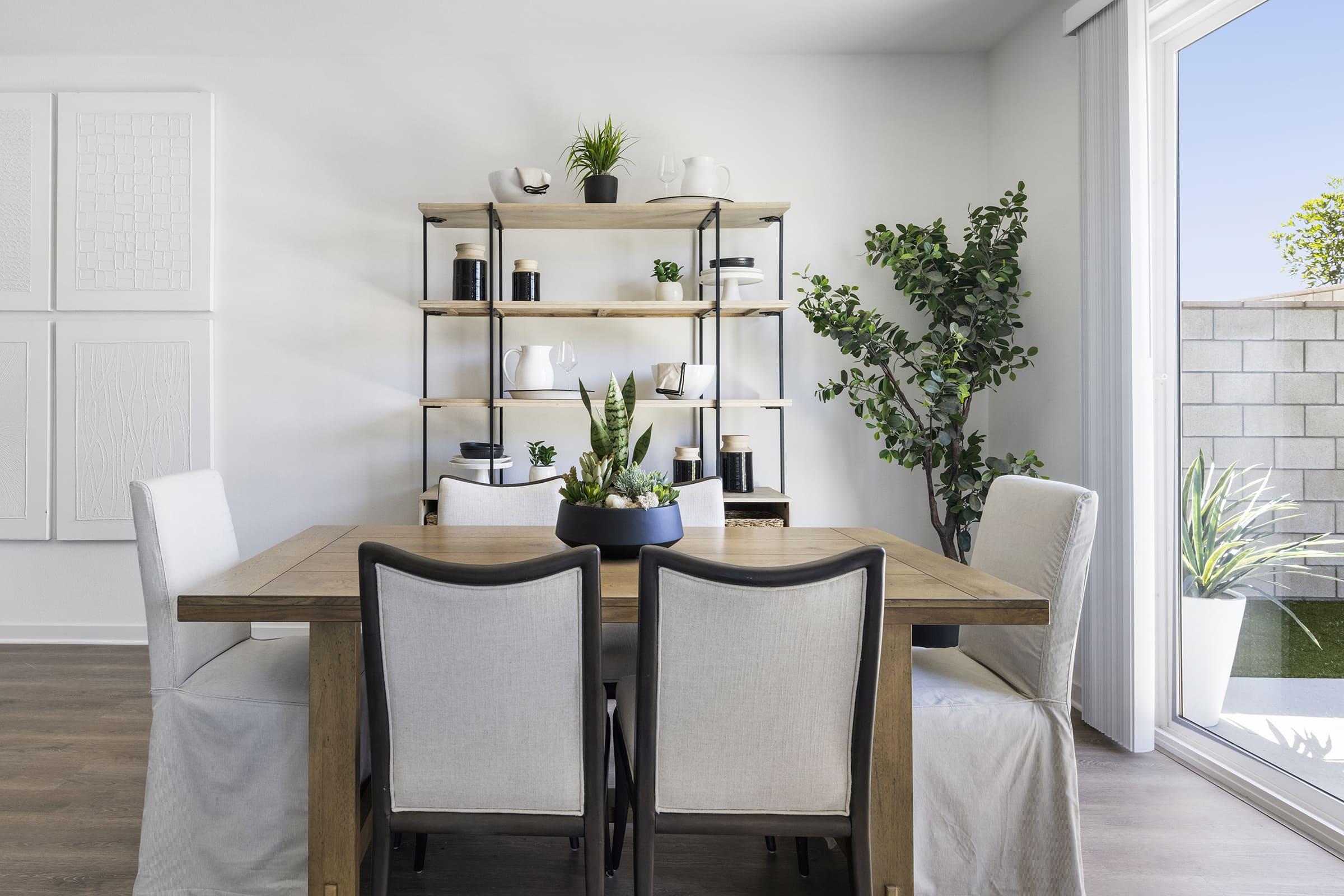 A modern dining area featuring a wooden table surrounded by four upholstered chairs. Behind the table is a tall bookshelf adorned with decorative plants, ceramic items, and black containers. Large windows allow natural light to fill the space, with greenery visible outside.