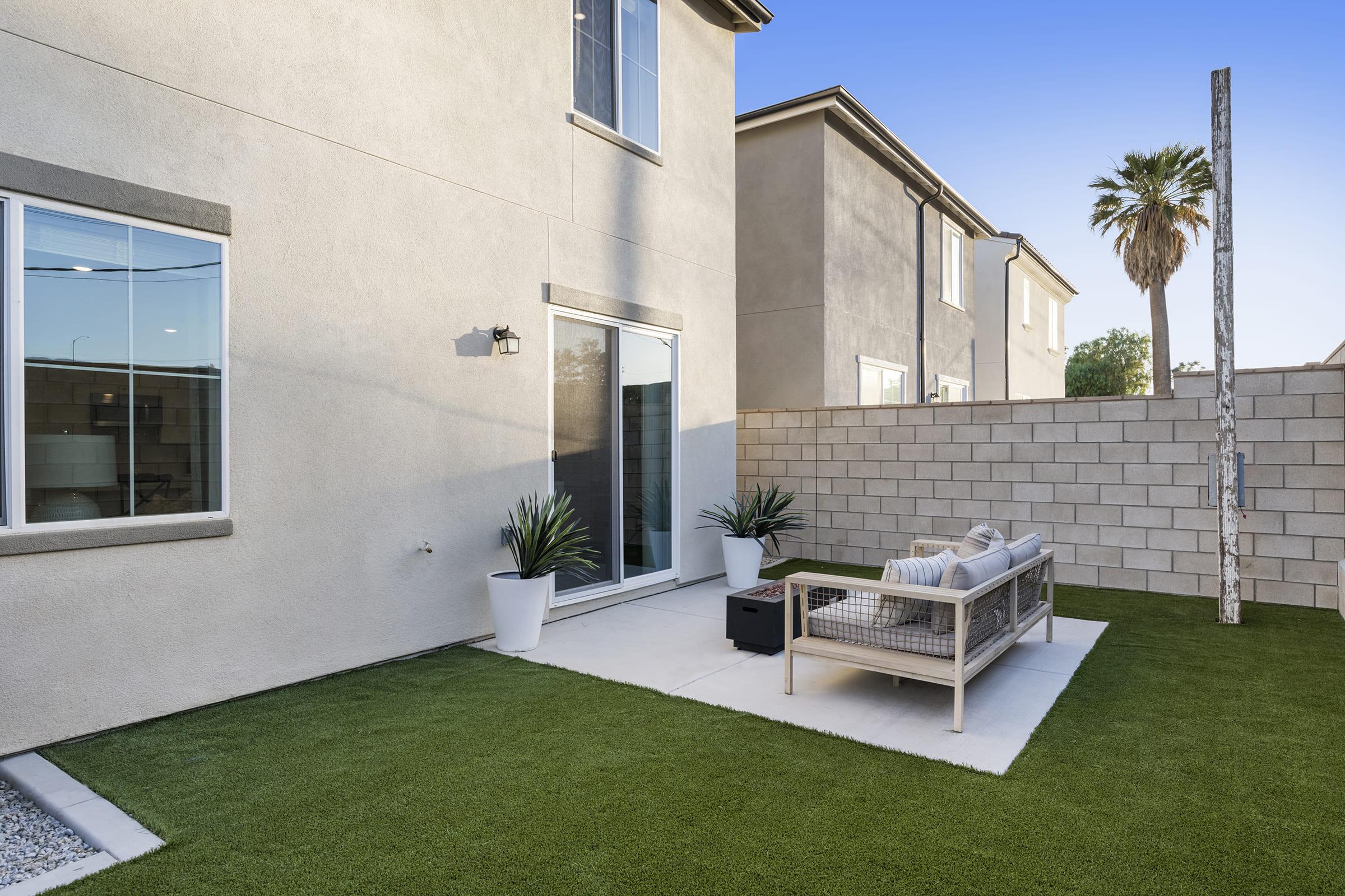 A modern backyard featuring a small patio area with a comfortable outdoor sofa and a coffee table. Lush artificial grass surrounds the seating area, and potted plants add greenery. The backdrop includes a light-colored house with large windows, and a palm tree is visible beyond the fence.