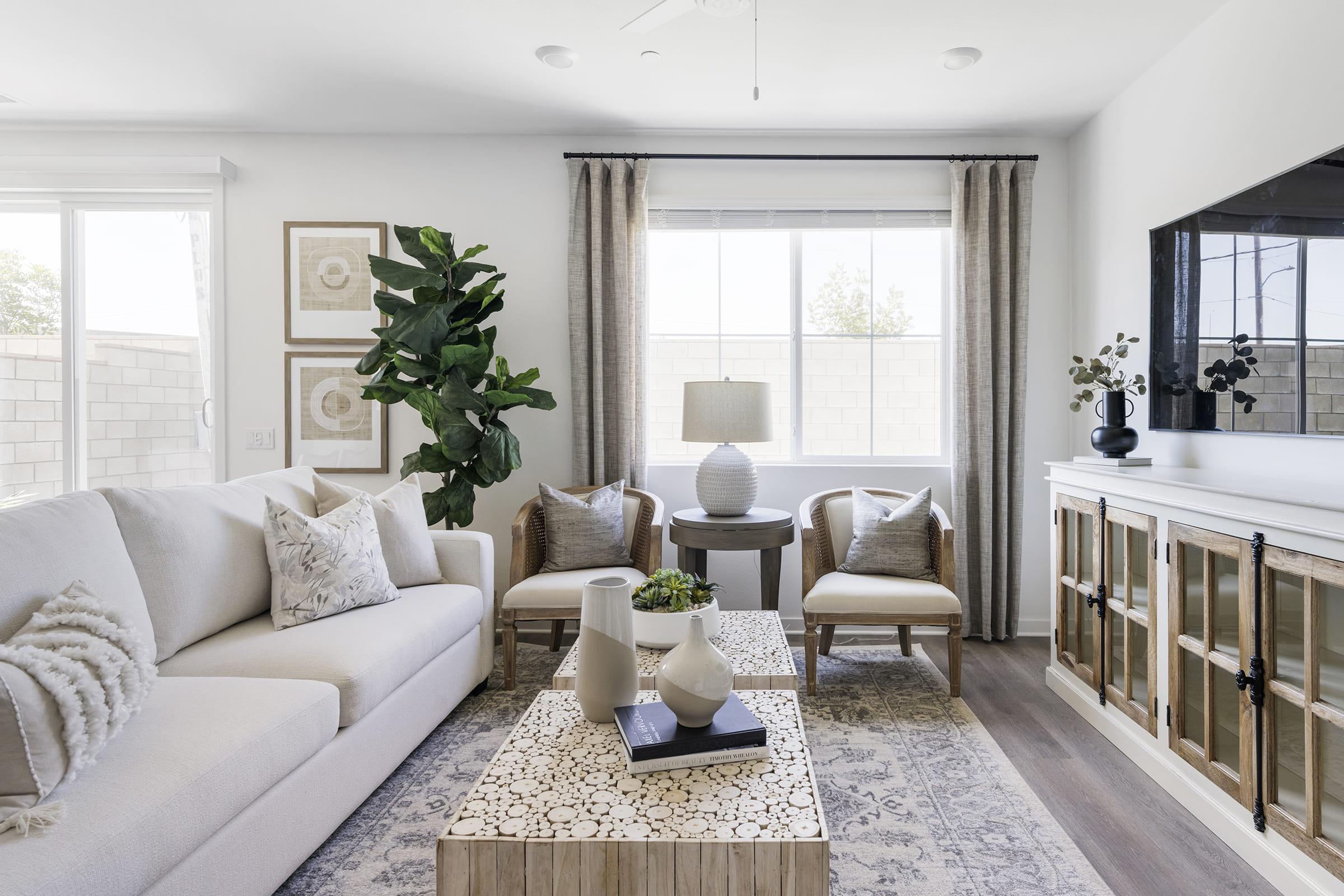 A modern living room featuring a light-colored couch with decorative pillows, a wooden coffee table, and a potted plant. There are two armchairs, a lamp on a side table, and framed art on the walls. Large windows brightening the space with neutral curtains and a stylish cabinet against the wall.