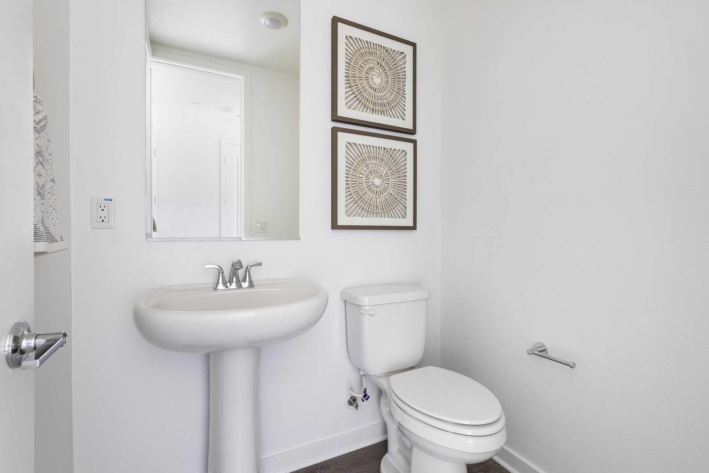 A clean, minimalist bathroom featuring a white toilet and a pedestal sink. There are two decorative framed pieces of art on the wall, and a mirror above the sink. The walls and floor have a neutral color scheme, contributing to a bright and airy atmosphere.