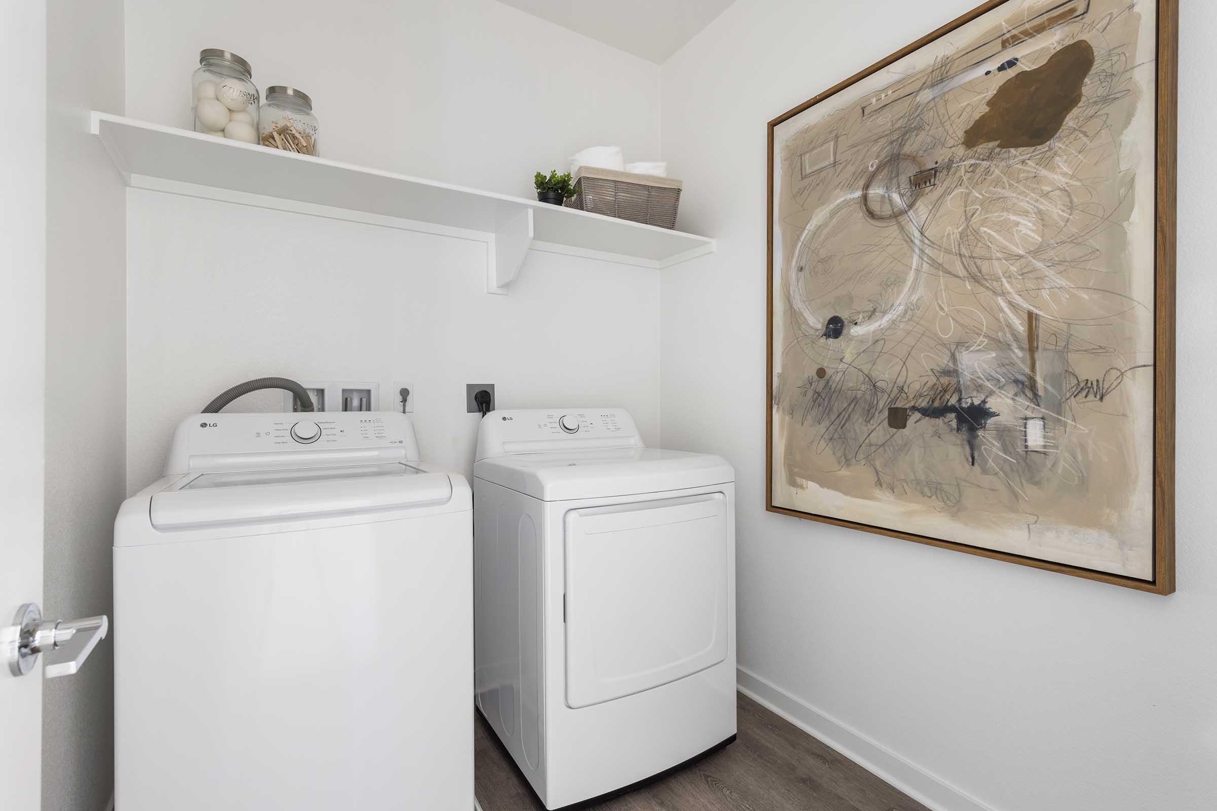 A clean laundry room featuring a stacked washer and dryer, with a shelf above holding storage containers. A framed abstract painting is hung on the wall, adding a modern touch to the space. The floors are light-colored, enhancing the bright and airy atmosphere of the room.