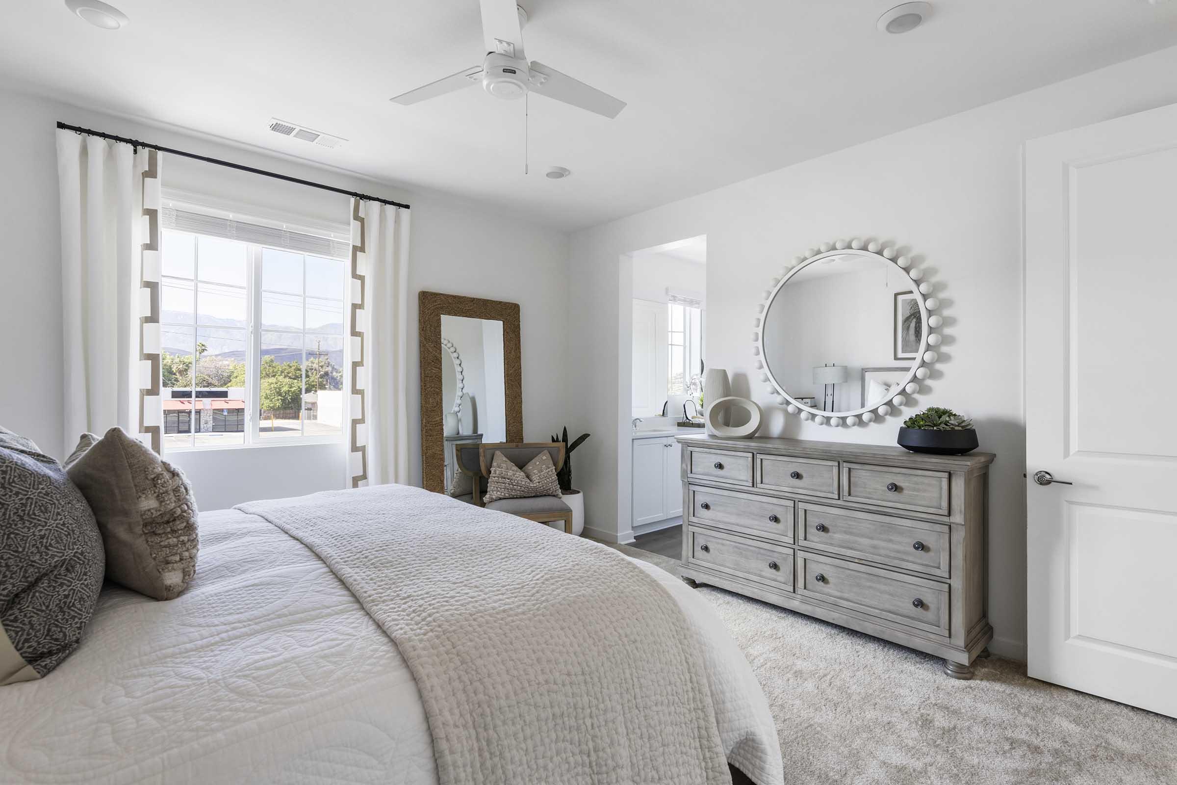 A bright and airy bedroom featuring a large window with sheer curtains, a comfortable bed with decorative pillows, a gray dresser, a round mirror above it, and a stylish chair. The room has soft neutral tones and a cozy atmosphere, complemented by a small plant and a large mirror reflecting the light.