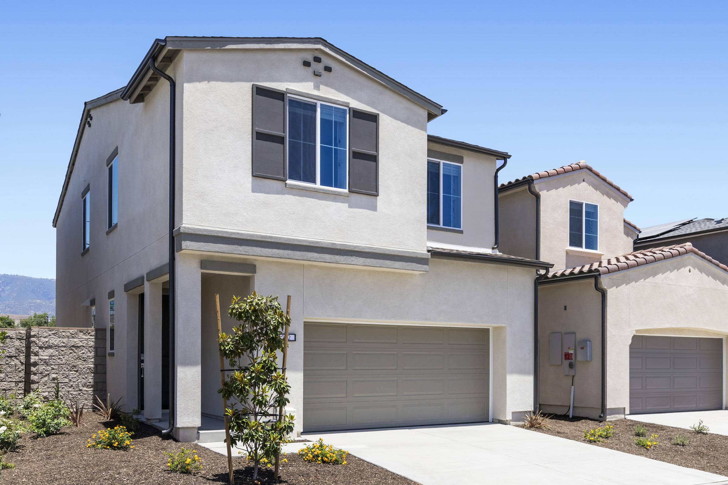 A modern two-story house with a light beige exterior, featuring a double garage, large windows, and decorative shutters. The front yard has minimal landscaping, including small shrubs and a tree. The backdrop shows clear blue skies and distant mountains.