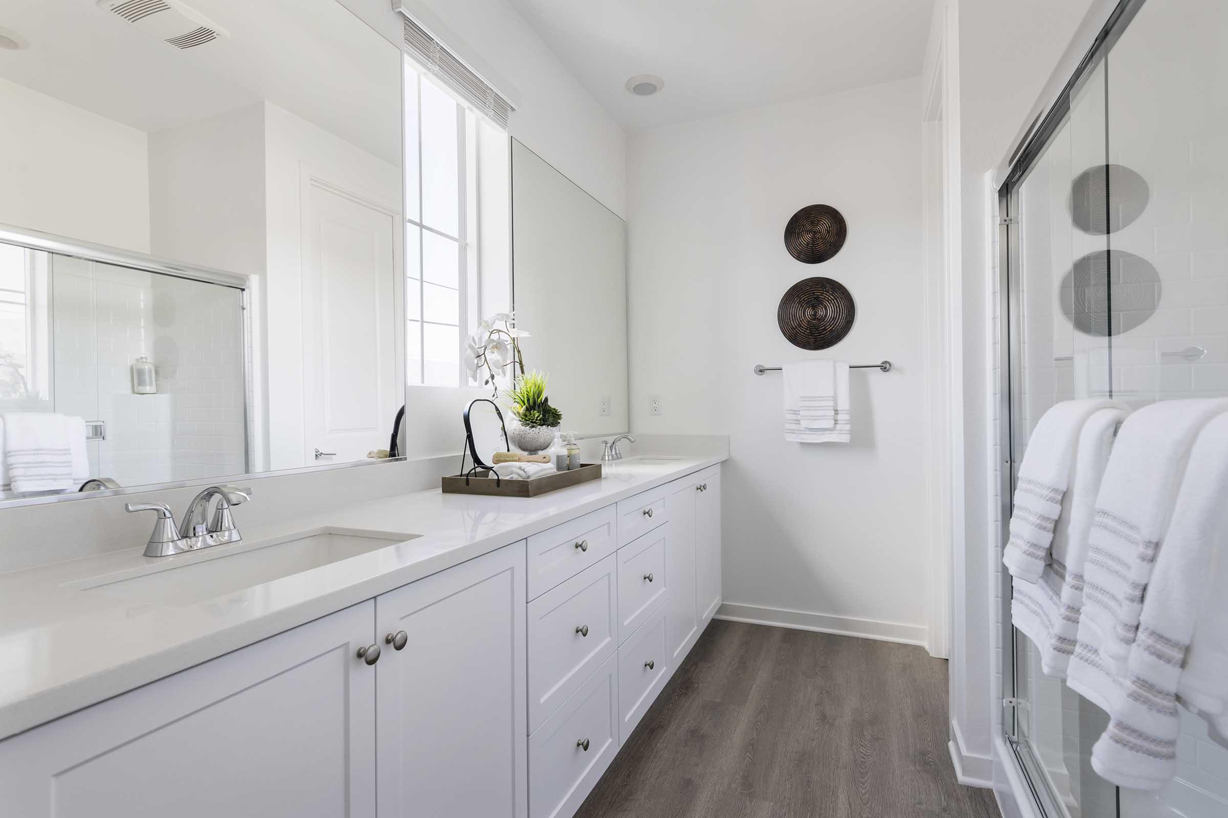 A modern bathroom featuring a double sink vanity with white cabinetry, a large mirror, and decorative wall plates. The space is bright and airy, with natural light coming through a window. White towels are neatly hung, and there is a plant displayed on the countertop, adding a touch of greenery.