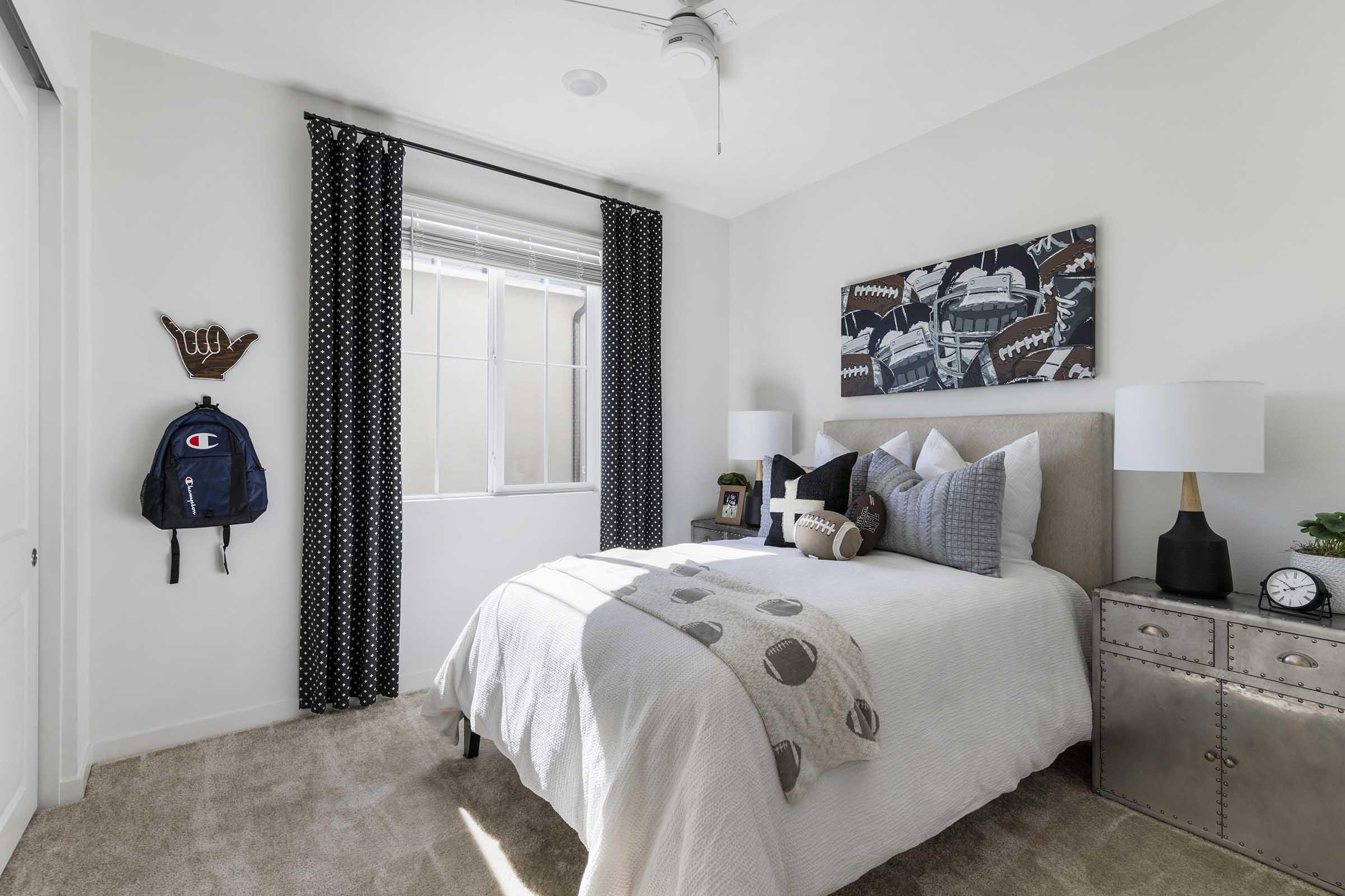 A cozy bedroom featuring a neatly made bed with gray and white bedding, pillows, and a decorative throw. There's a small nightstand with a clock and lamp beside the bed. A window with light curtains lets in natural light, while black and white curtains frame the window. A backpack hangs on the wall, adding a casual touch.