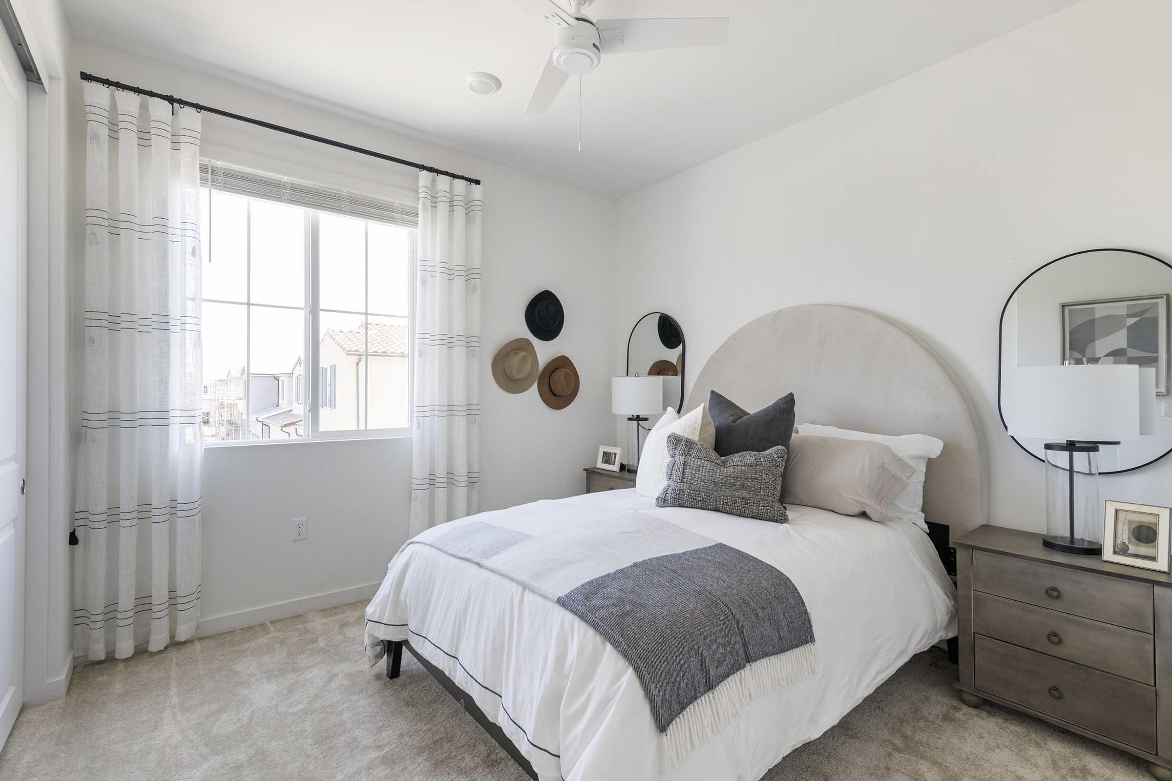 A bright and airy bedroom featuring a queen-sized bed with white bedding and gray accents. A round mirror is mounted above the headboard, complemented by stylish lamps on either side of a wooden nightstand. Decorated with wall hats and sheer curtains, the room has a neutral color palette and soft carpet.