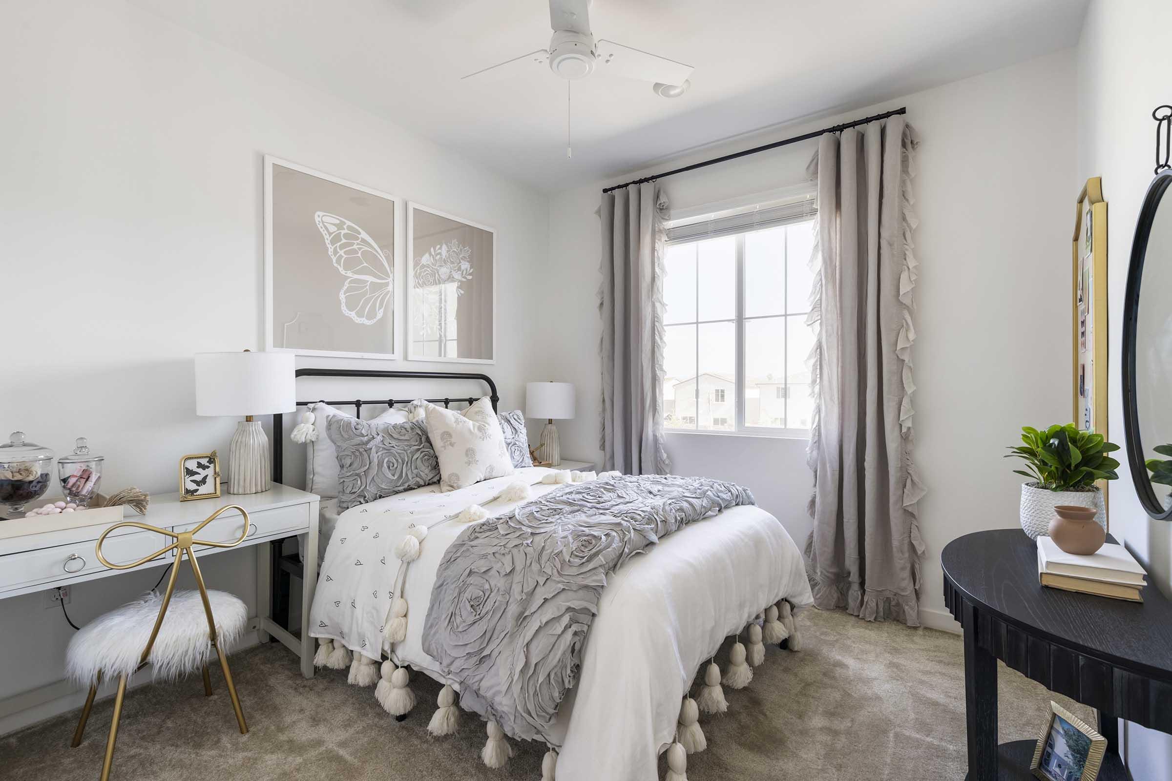 Cozy bedroom featuring a neatly made bed with a light gray, textured blanket adorned with pom-poms. There is a decorative headboard, stylish lamps on bedside tables, and a wall with butterfly artwork. A desk with a chair and a small plant adds to the inviting atmosphere, complemented by soft gray curtains and plush carpet.