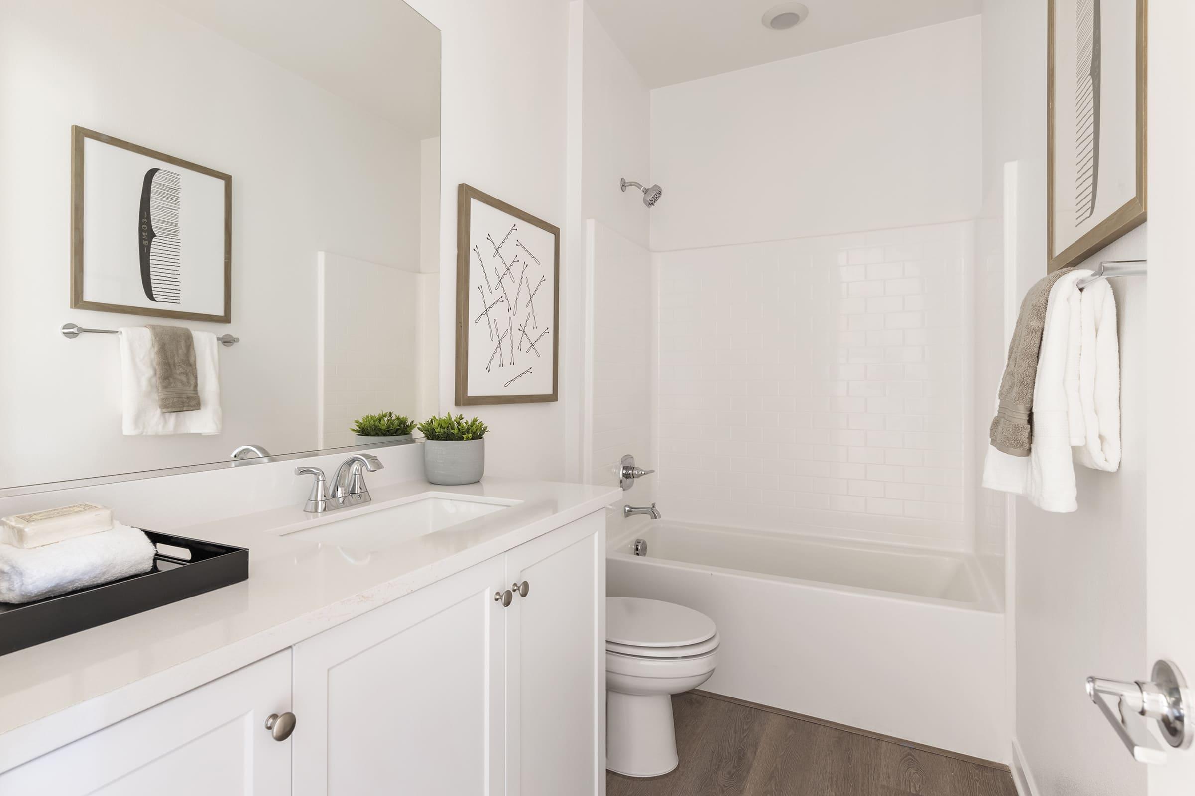 A modern bathroom featuring a clean design with white cabinetry, a rectangular mirror, and a simple framed artwork on the wall. The bathtub is white and accompanied by a chrome showerhead. A small plant and neatly arranged towels add a touch of decor on the countertop. The flooring is a light wood finish.