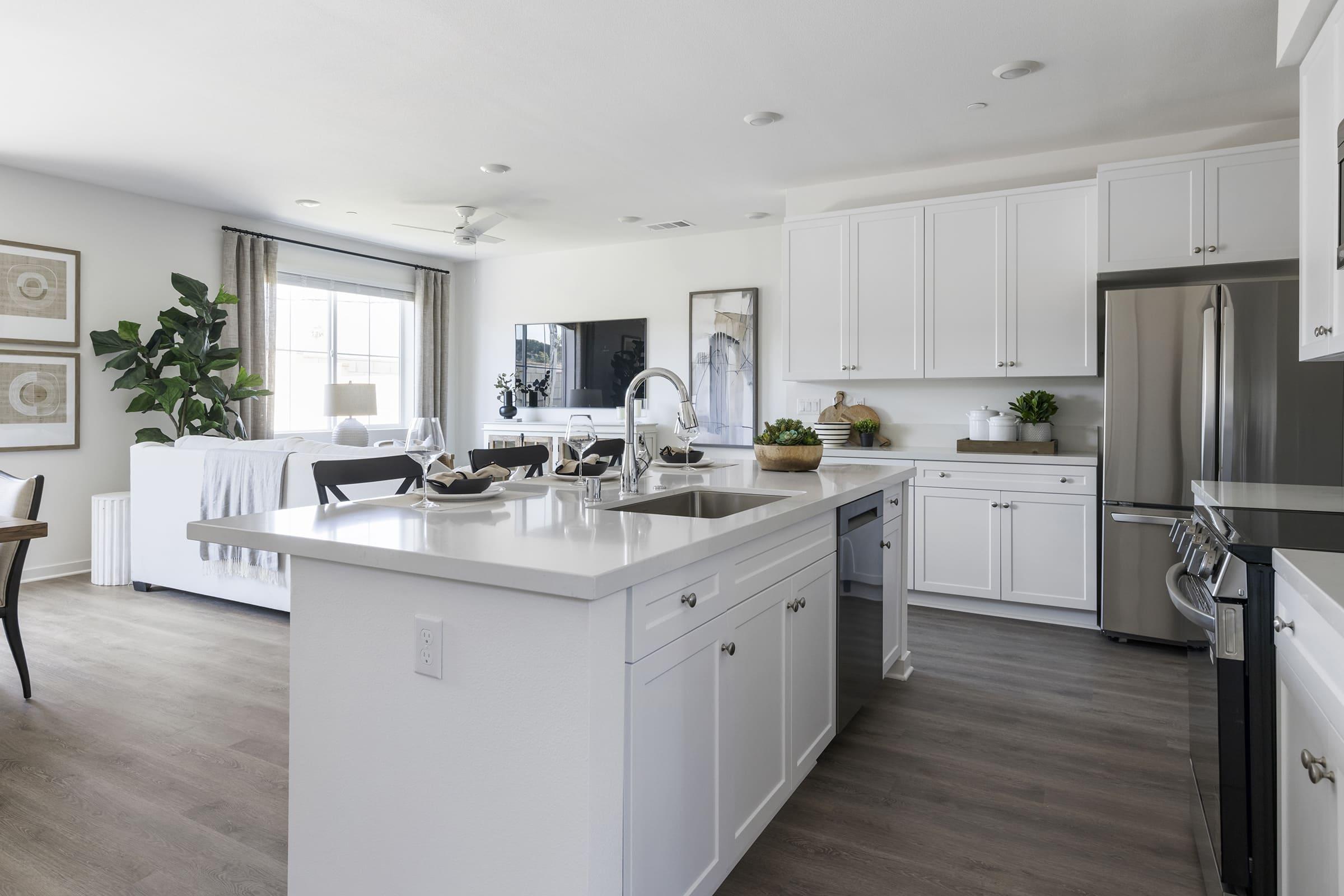 A modern kitchen featuring white cabinets, a large island with a sink, and stainless steel appliances. Bright and airy, the space includes a dining area with dark wooden chairs and a living area visible in the background, decorated with plants and contemporary furnishings. Natural light floods the room through large windows.