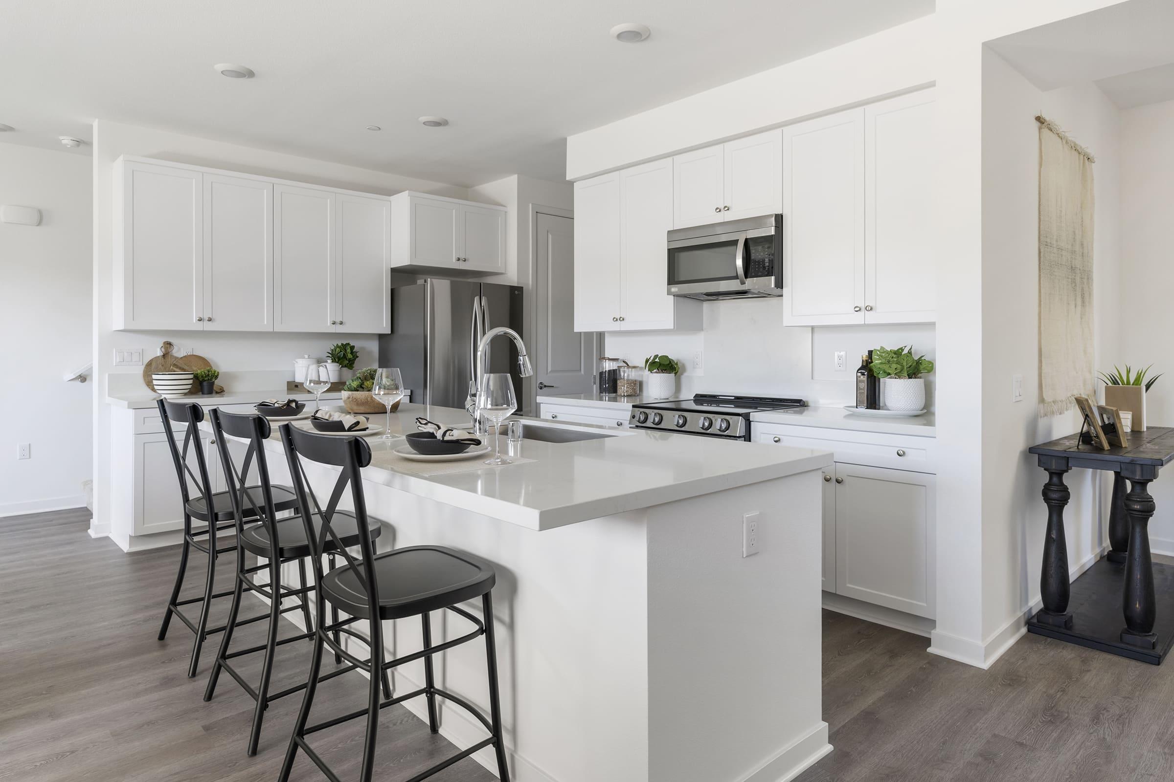 A modern kitchen featuring white cabinetry, stainless steel appliances, and a large island with bar stools. The kitchen is bright and open, with plants decor and a neutral color palette, creating a fresh and inviting atmosphere.