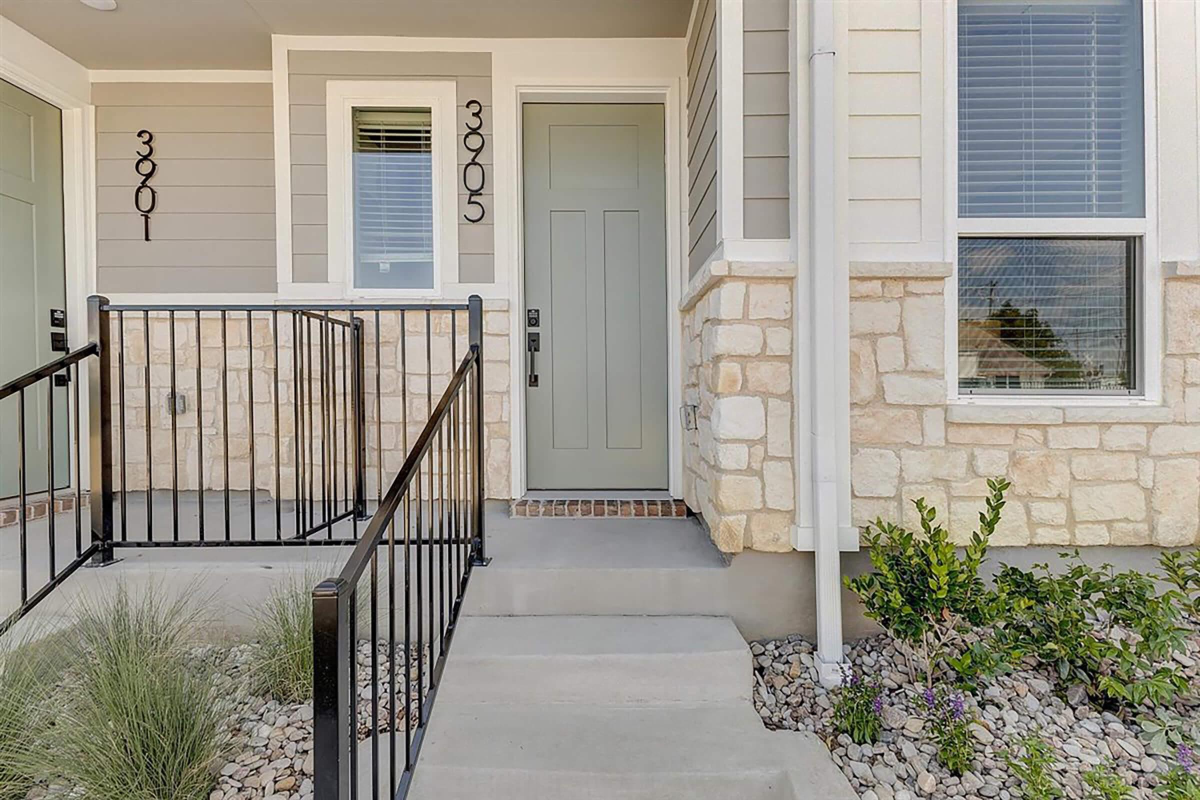 Front entrance of a modern two-story home featuring a gray door, stone and siding exterior, and a small pathway. Metal railings line the stairs, and there are decorative plants and rocks nearby. Windows are visible on the sides, providing additional light.