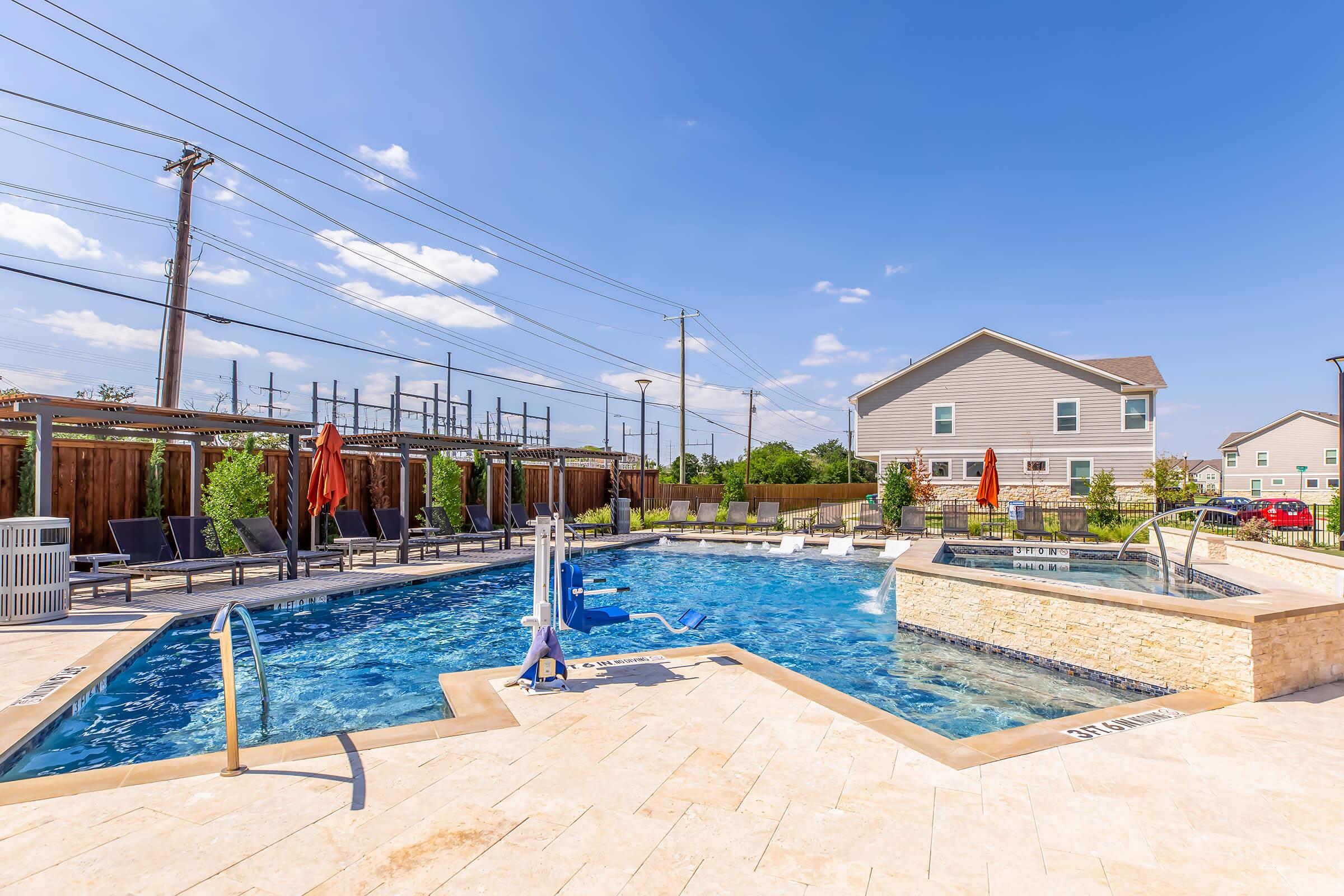 A bright outdoor pool area featuring a clear blue pool with lounge chairs along the edges. There is a hot tub beside the pool, surrounded by vibrant greenery. Orange umbrellas provide shade, and nearby are utility poles and a residential building in the background under a clear blue sky.
