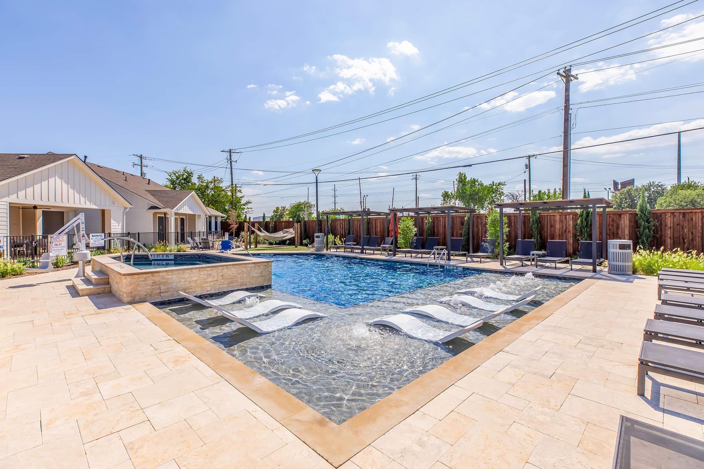 A bright outdoor swimming pool area featuring a large pool with lounge chairs and a spa. Surrounding the pool are wooden fences and a sunny blue sky with a few clouds. The area is landscaped with greenery, creating a relaxing atmosphere for leisure and enjoyment.