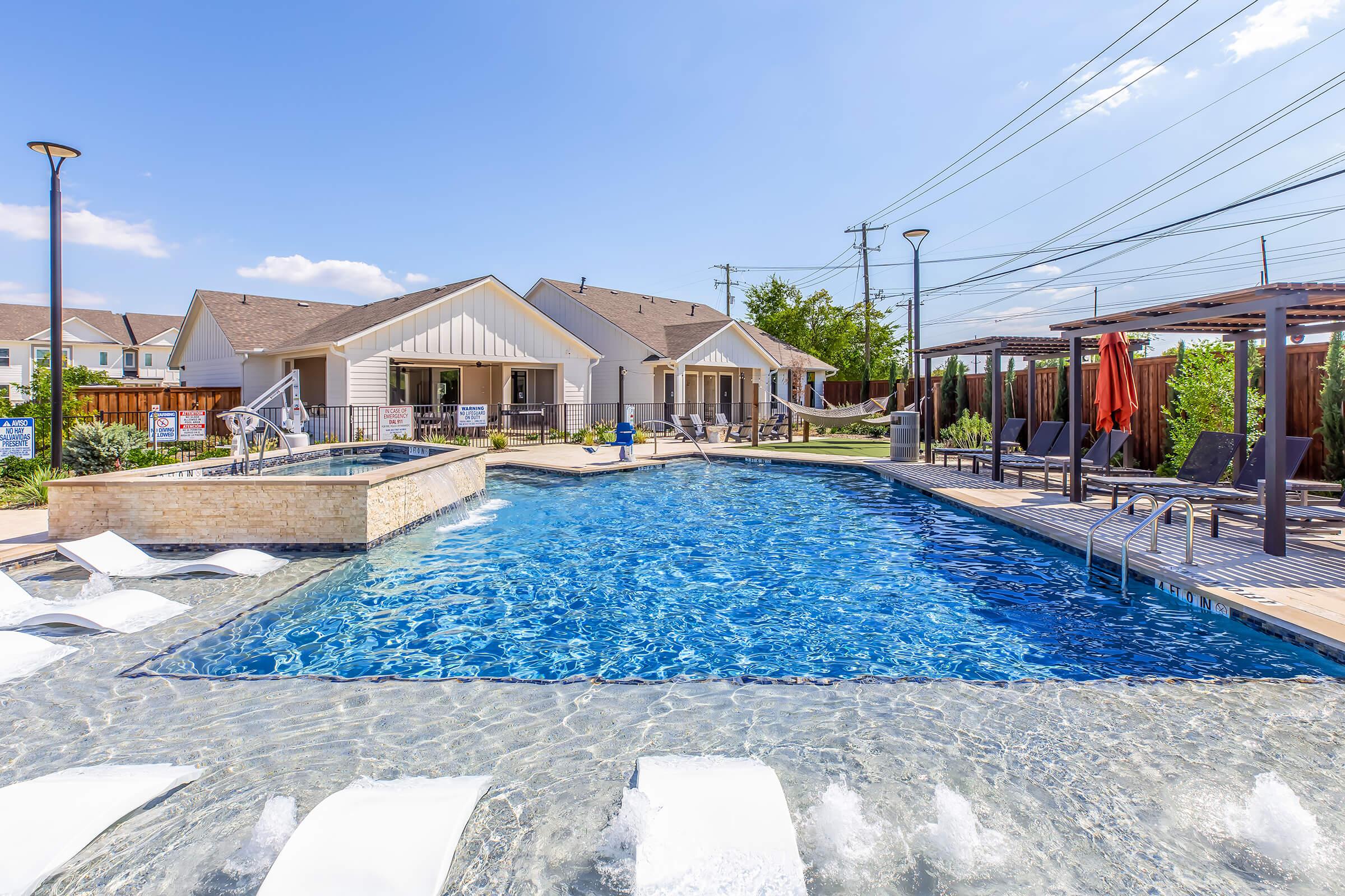 A vibrant outdoor swimming pool area featuring a clear blue pool, lounge chairs, and shaded cabanas. Surrounding the pool are well-maintained buildings, green landscaping, and a sunny sky with a few clouds, creating a relaxing and inviting atmosphere for leisure activities.