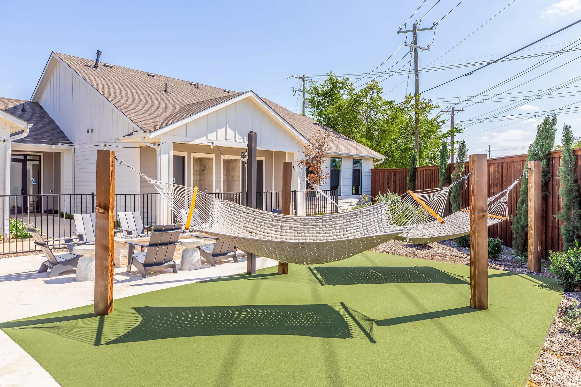 A cozy outdoor space featuring a hammock suspended between two wooden posts on a green lawn. In the background, there's a modern house with a porch and large windows, bordered by a wooden fence and tall plants. Bright blue skies and utility poles are visible, creating a relaxed, inviting atmosphere.