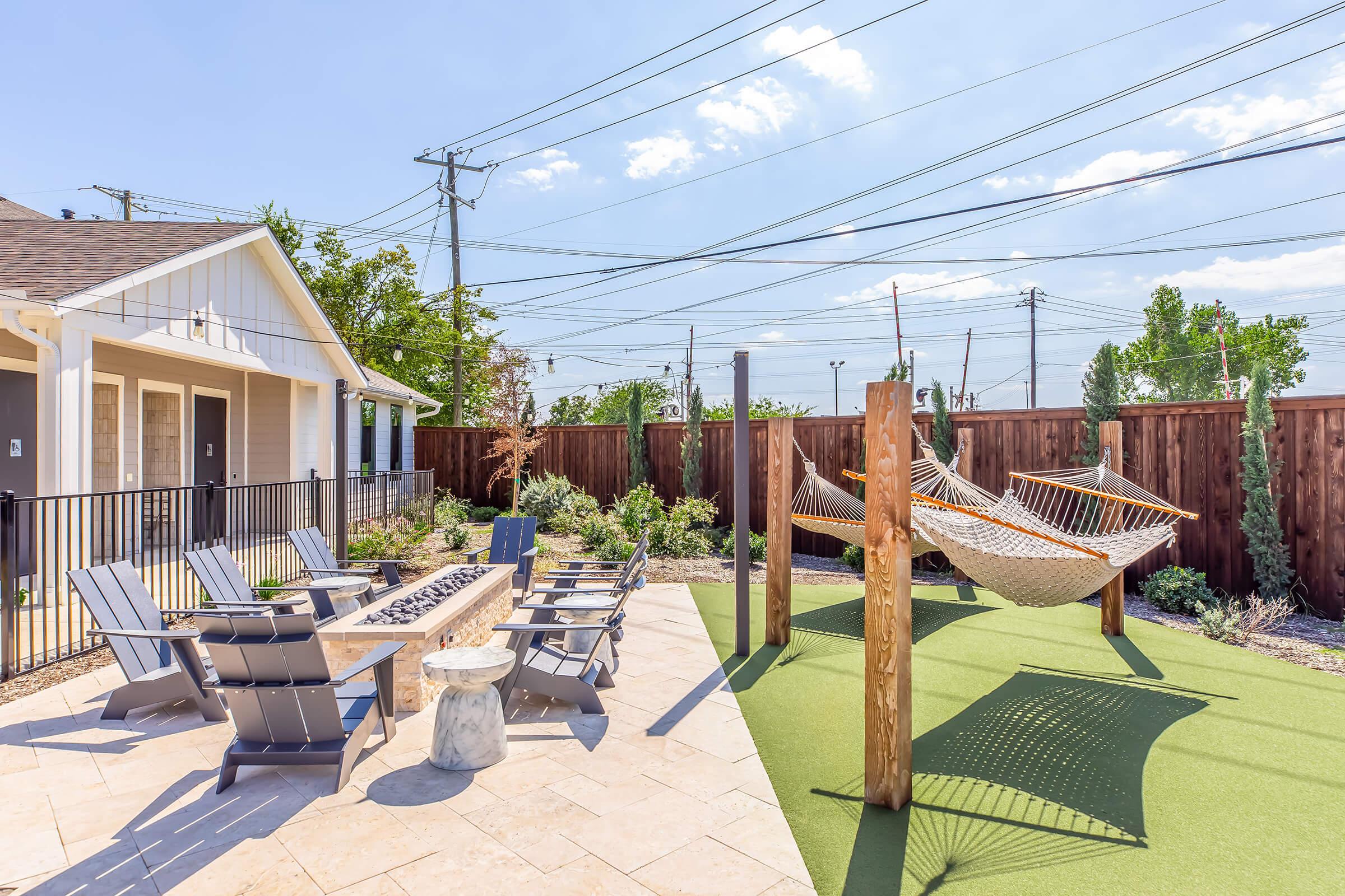Outdoor recreational area featuring a fire pit surrounded by lounge chairs, two hammocks on wooden supports, and turf flooring. The space is enclosed by a wooden fence and includes greenery, with a clear blue sky and power lines in the background. Ideal for relaxation and social gatherings.